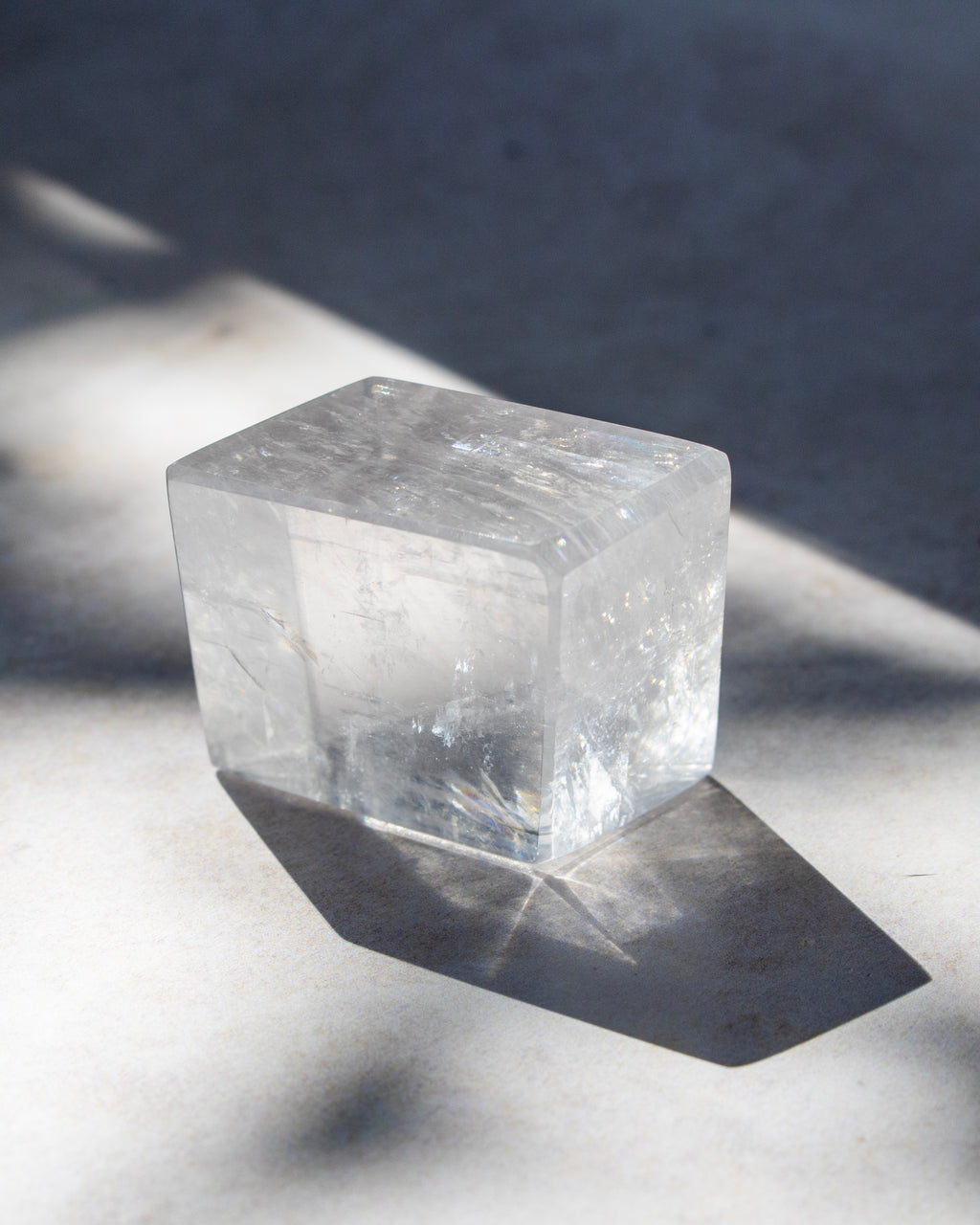 A clear, translucent cube-shaped crystal or stone block with natural internal inclusions, photographed on a light surface with strong diagonal shadows.