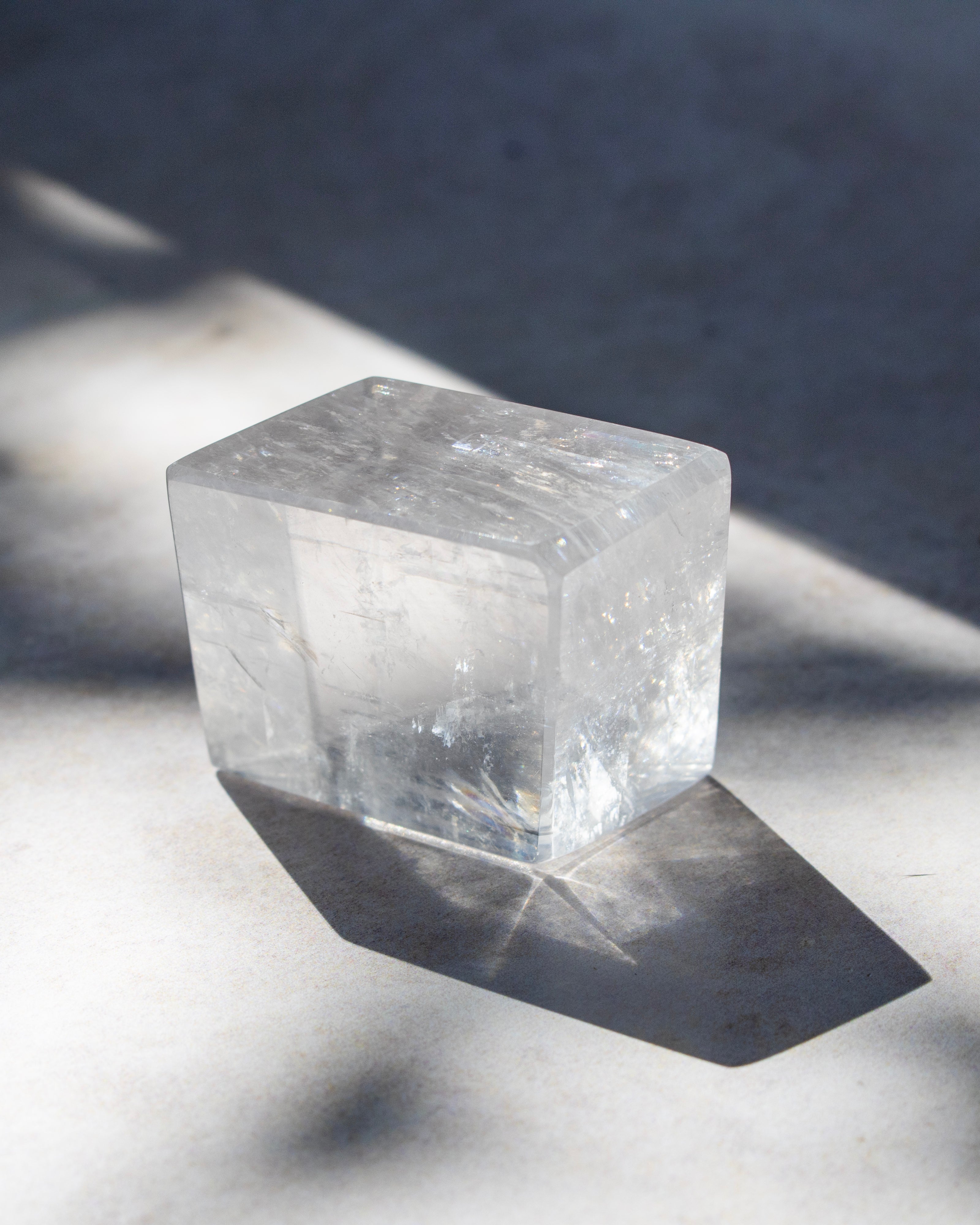 A clear, translucent cube-shaped crystal or stone block with natural internal inclusions, photographed on a light surface with strong diagonal shadows.