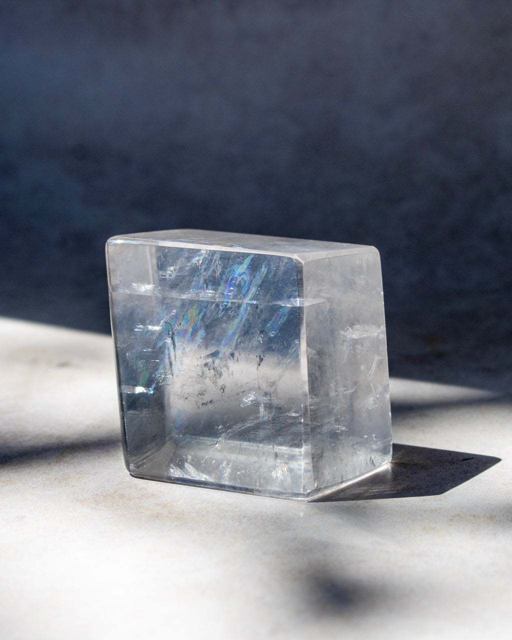 A clear rectangular crystal block with natural internal inclusions and subtle blue iridescence, photographed on a light surface with strong sunlight and shadow against a dark blue-gray background.