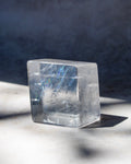 A clear rectangular crystal block with natural internal inclusions and subtle blue iridescence, photographed on a light surface with strong sunlight and shadow against a dark blue-gray background.