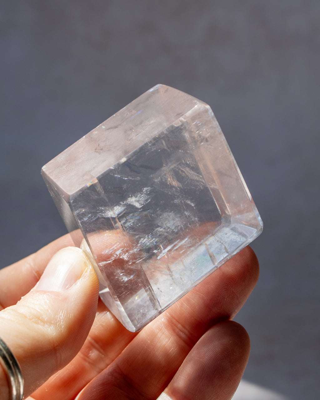 A hand holding a clear, faceted crystal cube with natural internal inclusions against a dark gray background.