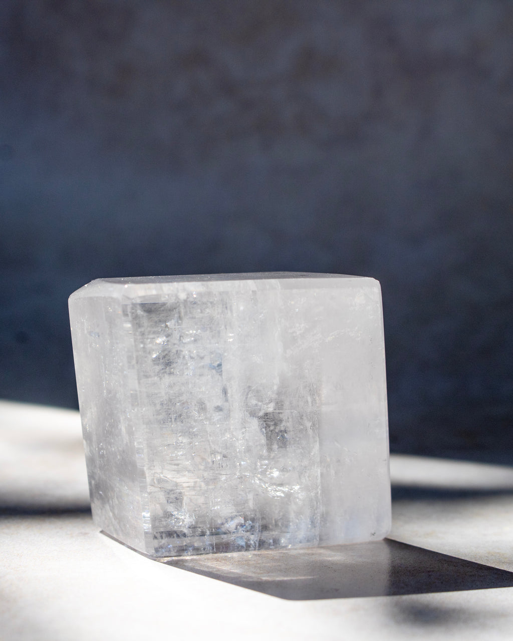 A clear, faceted crystal block with natural internal inclusions, photographed on a light surface with a dark textured background and a sharp shadow.
