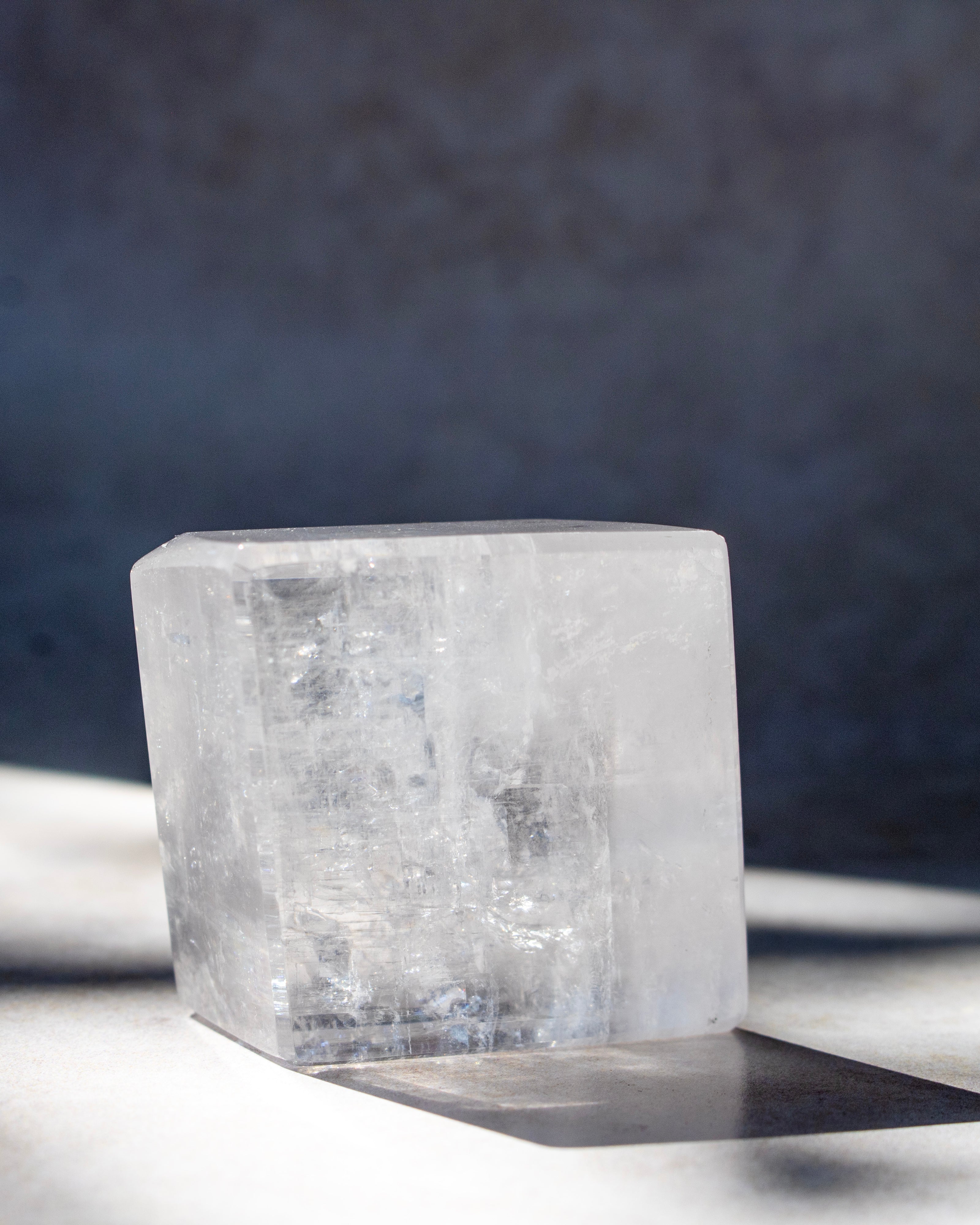 A clear, faceted crystal block with natural internal inclusions, photographed on a light surface with a dark textured background and a sharp shadow.