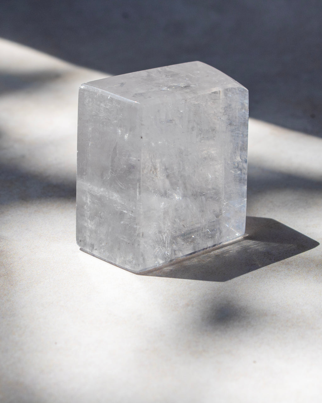 A clear rectangular crystal or stone block with rounded top corners, photographed on a light surface in strong natural sunlight with a sharp shadow.