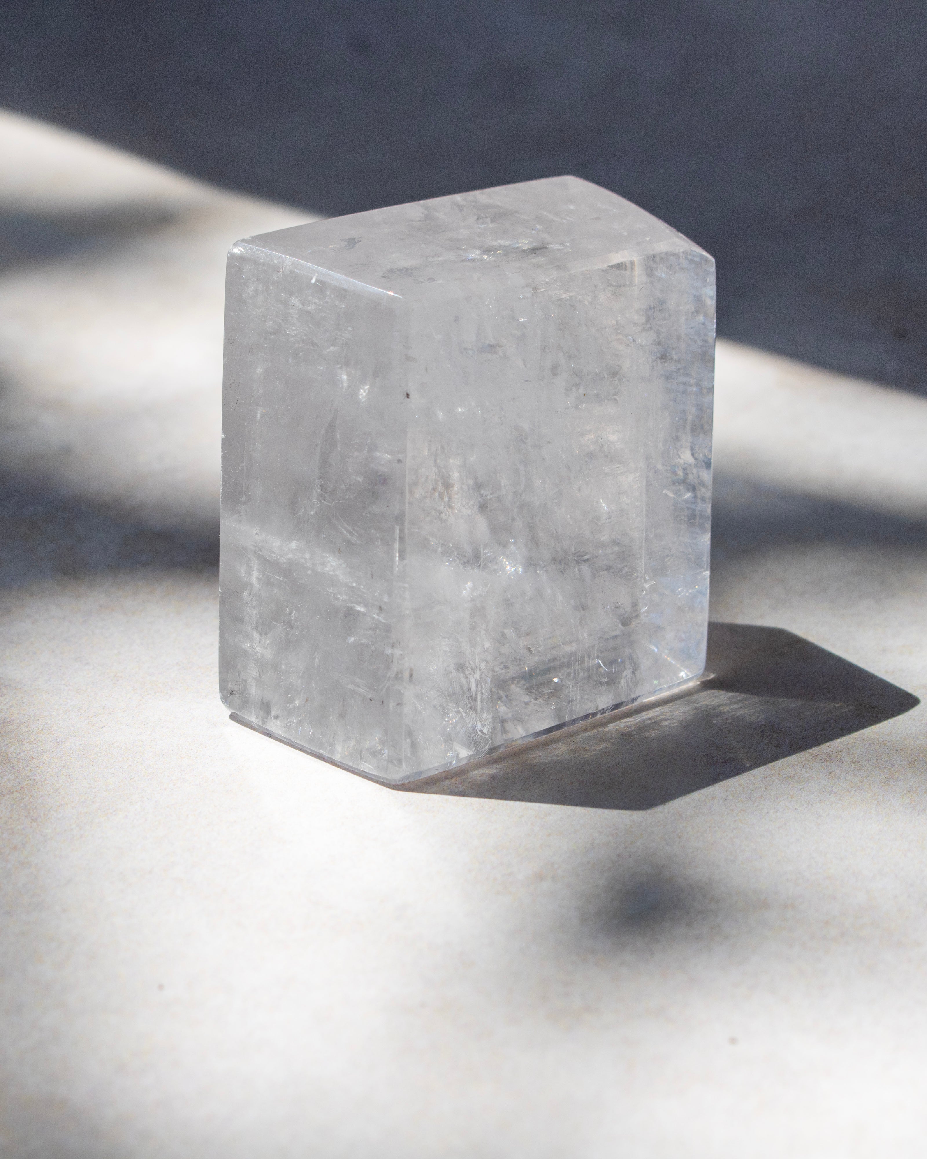 A clear rectangular crystal or stone block with rounded top corners, photographed on a light surface in strong natural sunlight with a sharp shadow.
