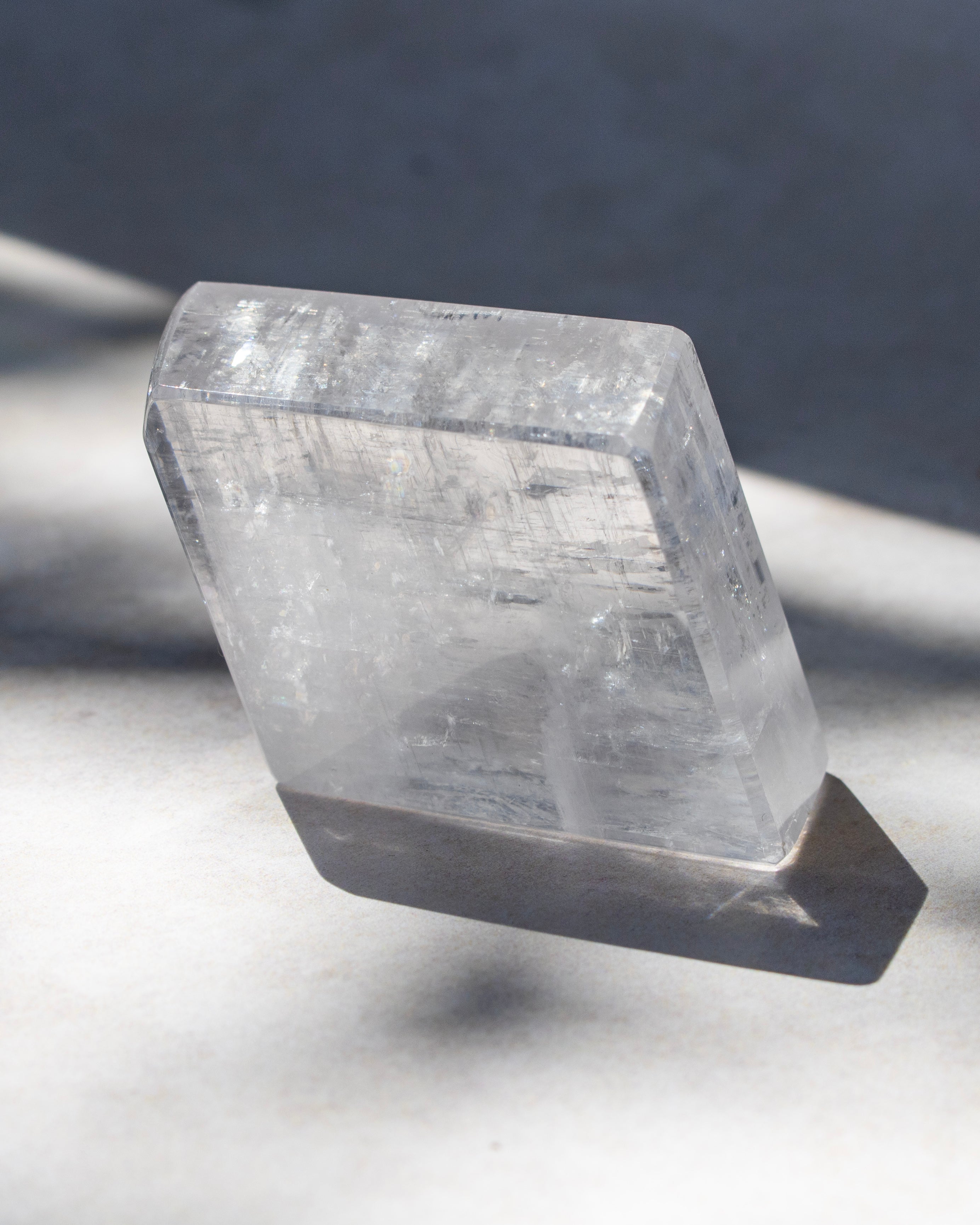 A clear, polished rectangular crystal or gemstone specimen displayed on a dark stand against a soft gray background with natural shadows.