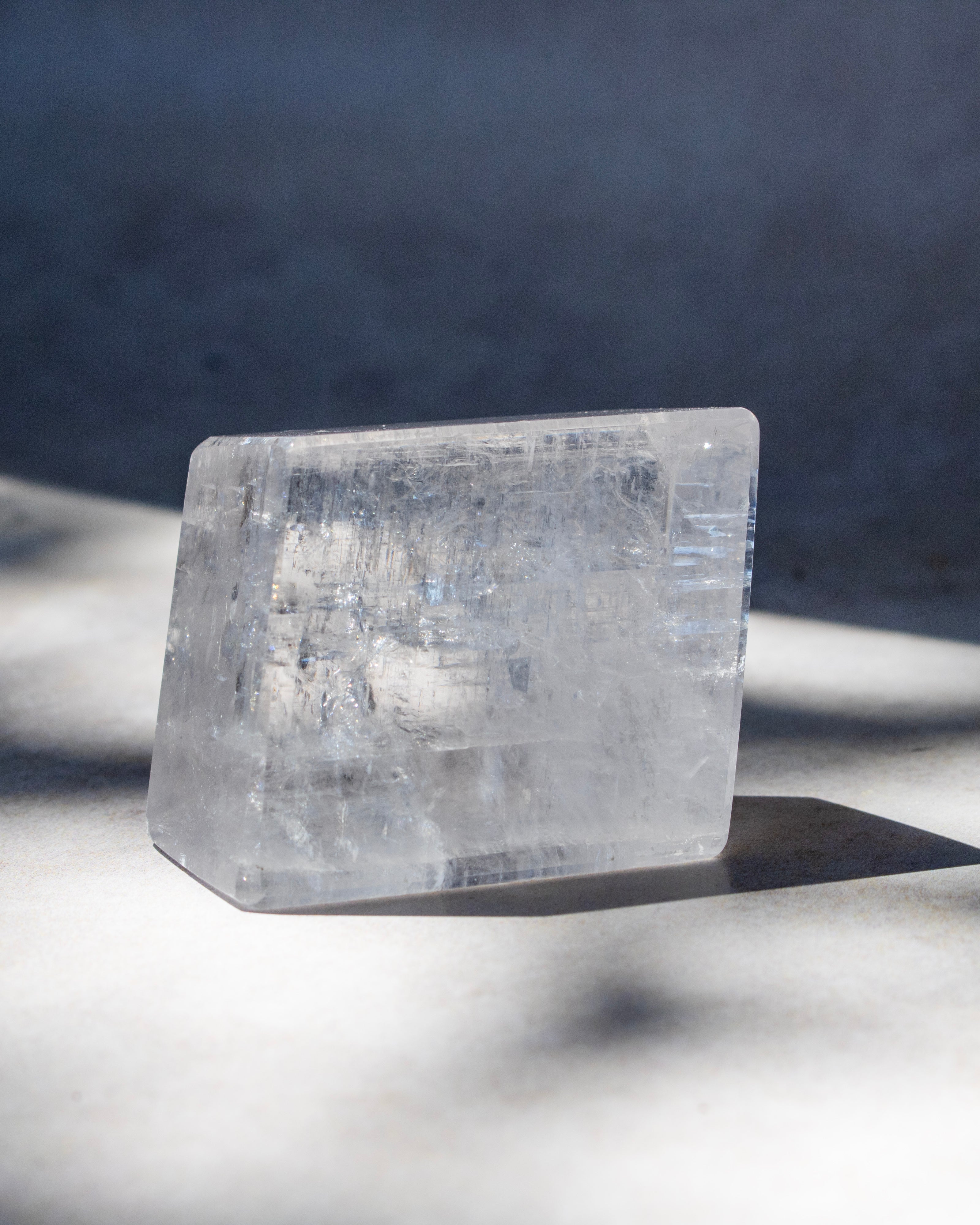 A rectangular clear crystal block with natural internal inclusions, standing on a light surface under direct sunlight with soft shadows.