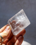 A hand holding a clear, geometric crystal or stone with visible internal striations against a soft gray background.