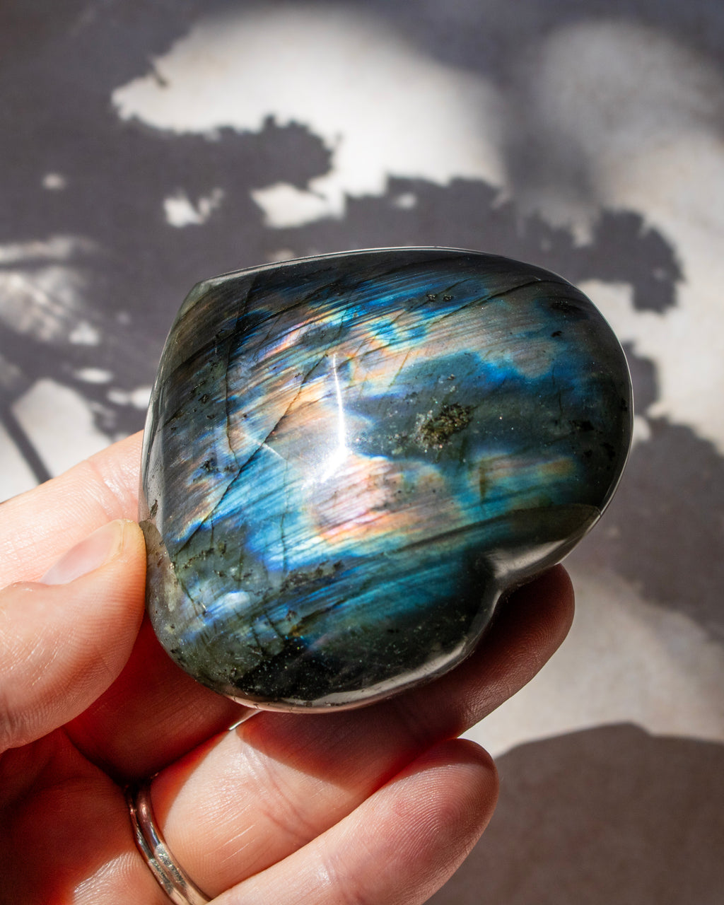 A polished labradorite stone held in a hand, showing a shimmering blue, green, and gold iridescent finish against a shadowed light background.