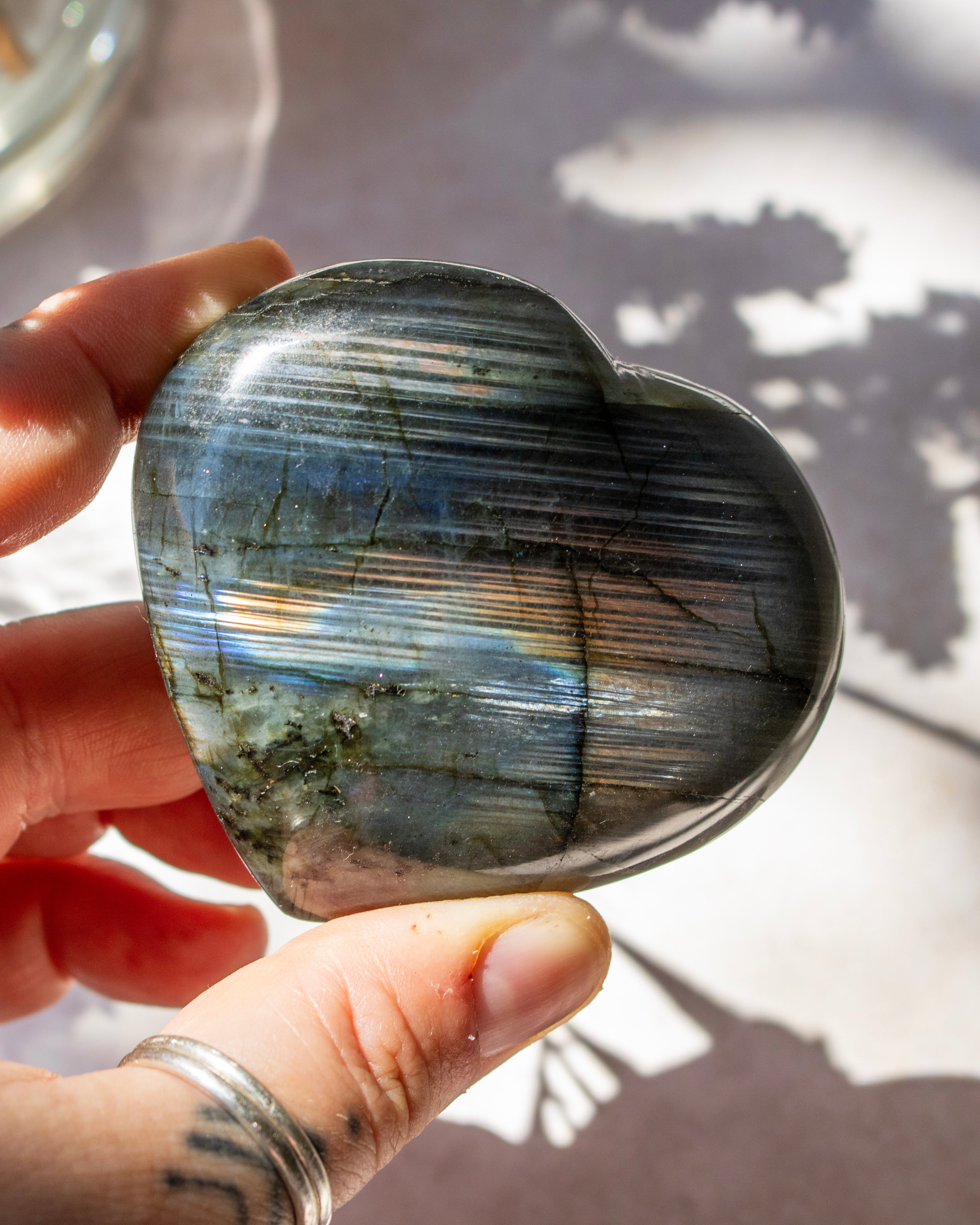 A polished heart-shaped labradorite stone held in a hand, showing its characteristic blue, gray, and iridescent rainbow sheen under bright sunlight.
