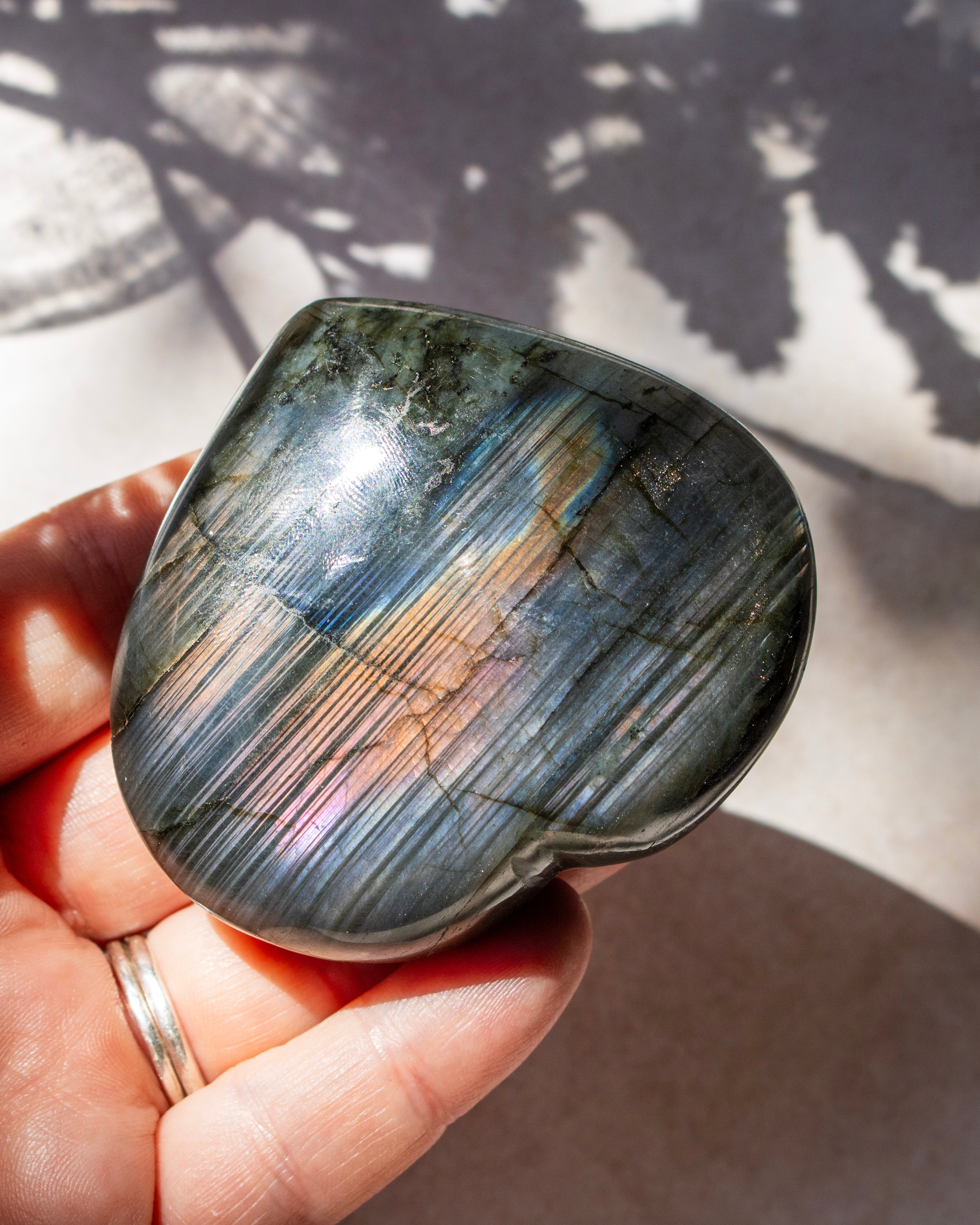 A polished labradorite stone held in a hand, showing a smooth rounded shape and natural banding with iridescent flashes of pink, blue, and gold under direct sunlight.