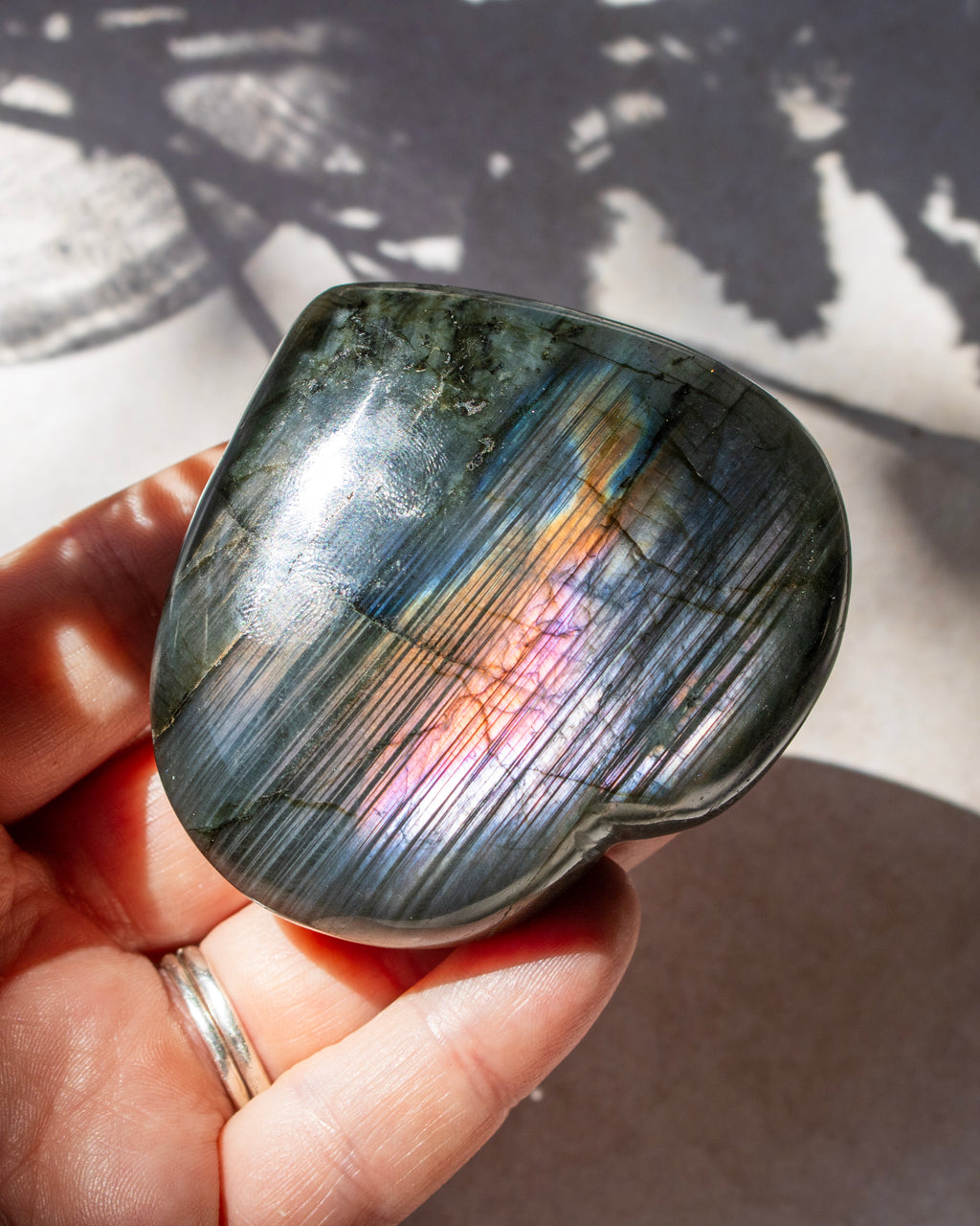 A polished tumbled labradorite stone held in a hand, showing a gray base with strong iridescent rainbow flashes under direct sunlight.
