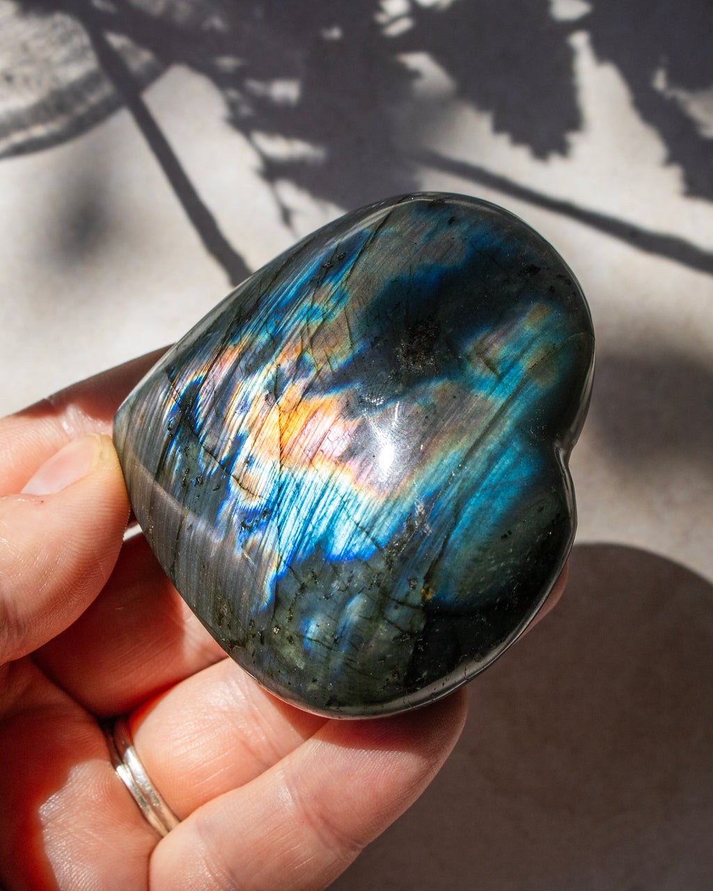 A heart-shaped labradorite stone held in a hand, showcasing its vibrant blue, green, and orange iridescent play-of-color against a sunlit background with leaf shadows.