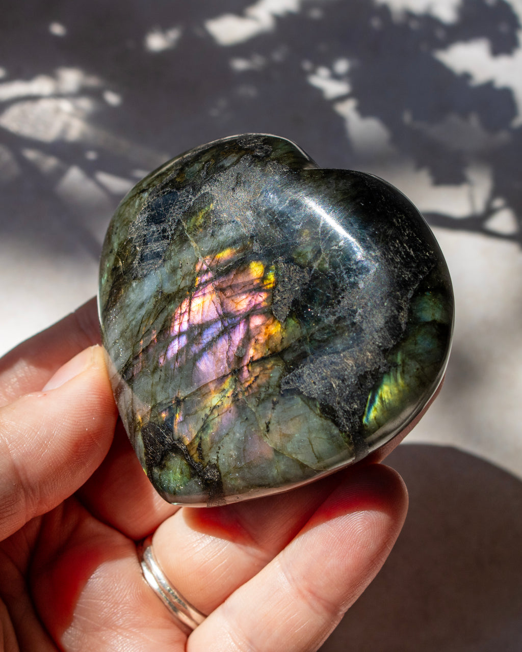 A polished heart-shaped labradorite stone held in a person's hand, showing a shimmering iridescent play-of-color with green, purple, and gold tones against a sunlit background with leaf shadows.
