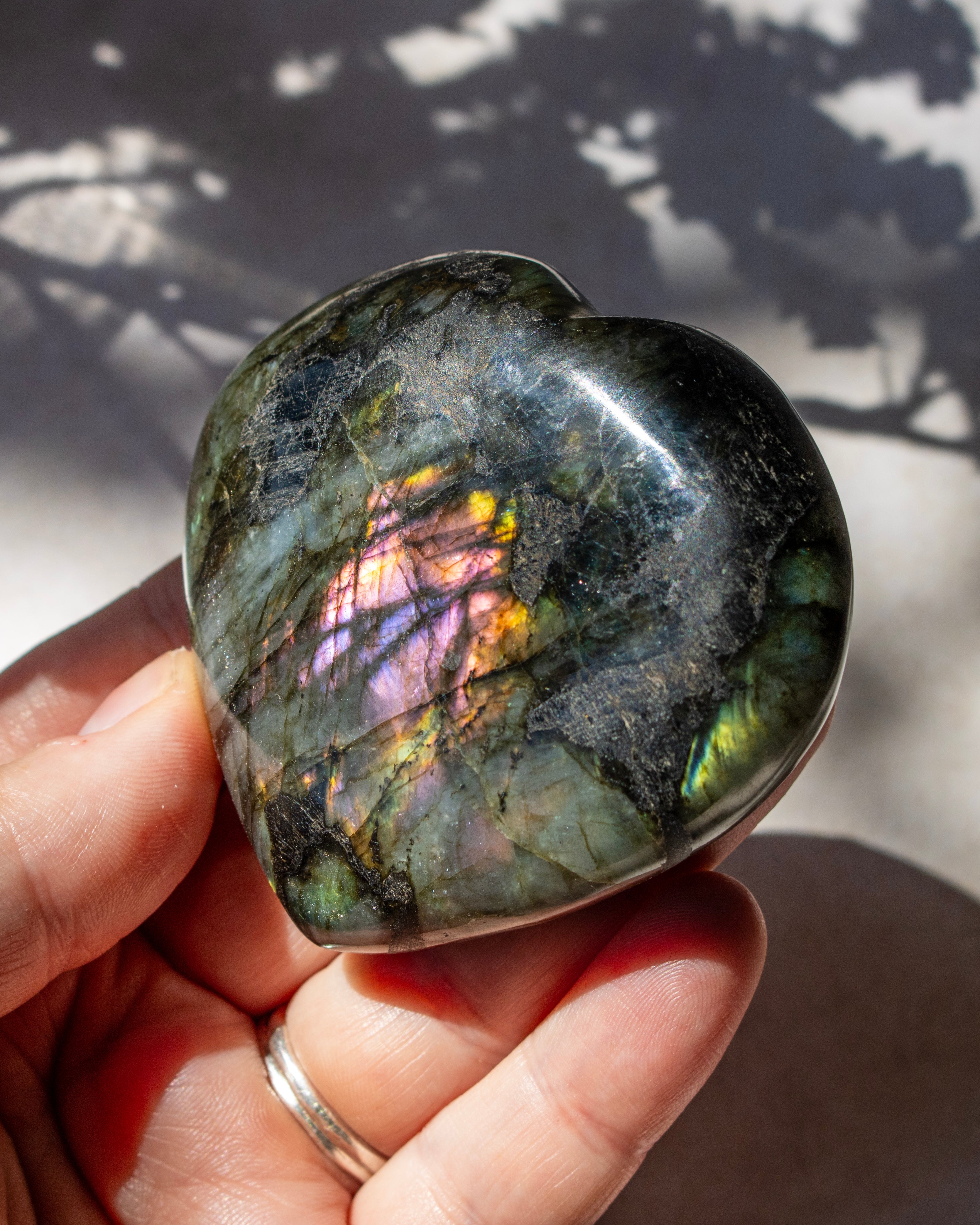 A polished heart-shaped labradorite stone held in a person's hand, showing a shimmering iridescent play-of-color with green, purple, and gold tones against a sunlit background with leaf shadows.