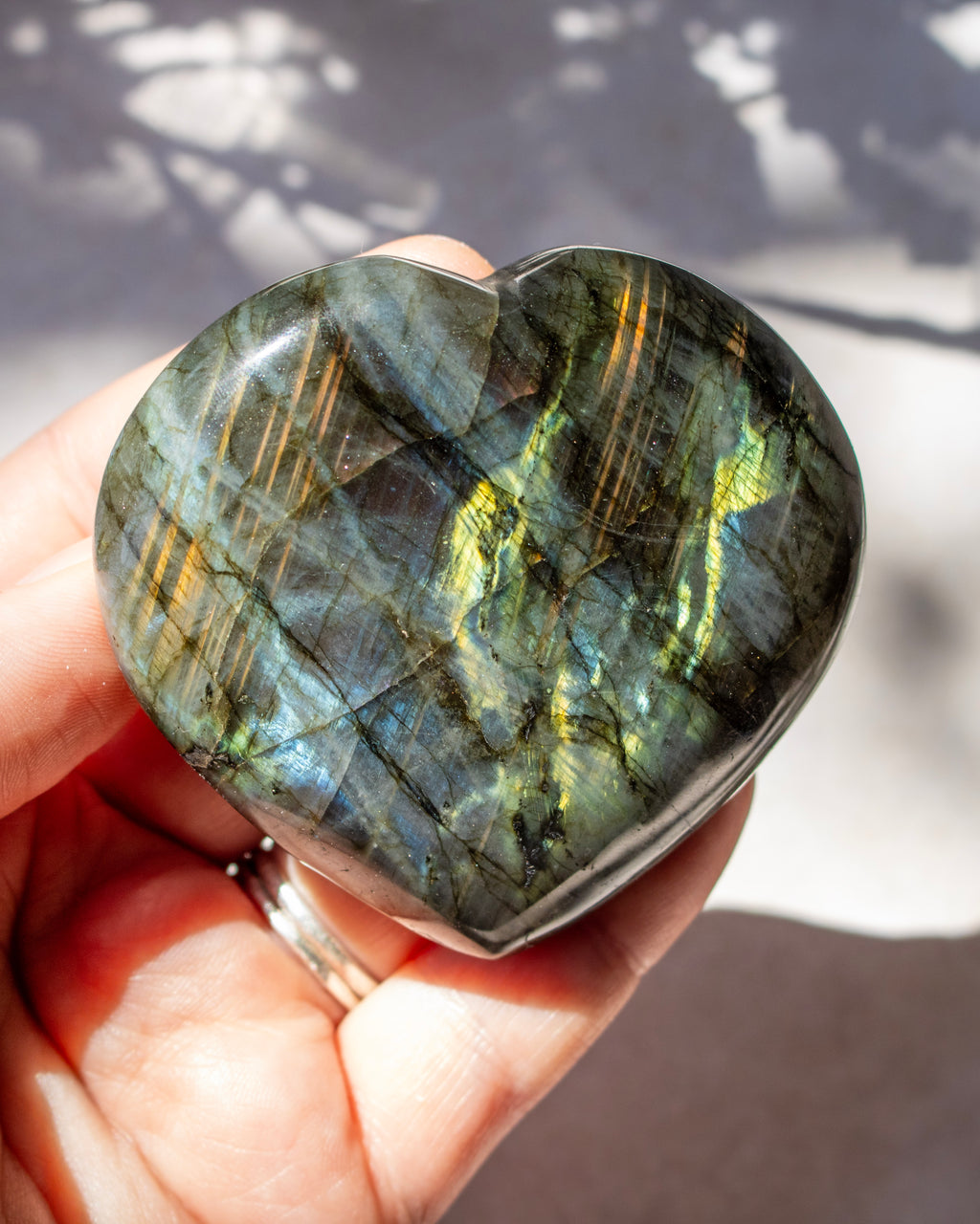 A heart-shaped polished labradorite stone held in a hand, showing shimmering green, gold, and blue flashes under natural sunlight.