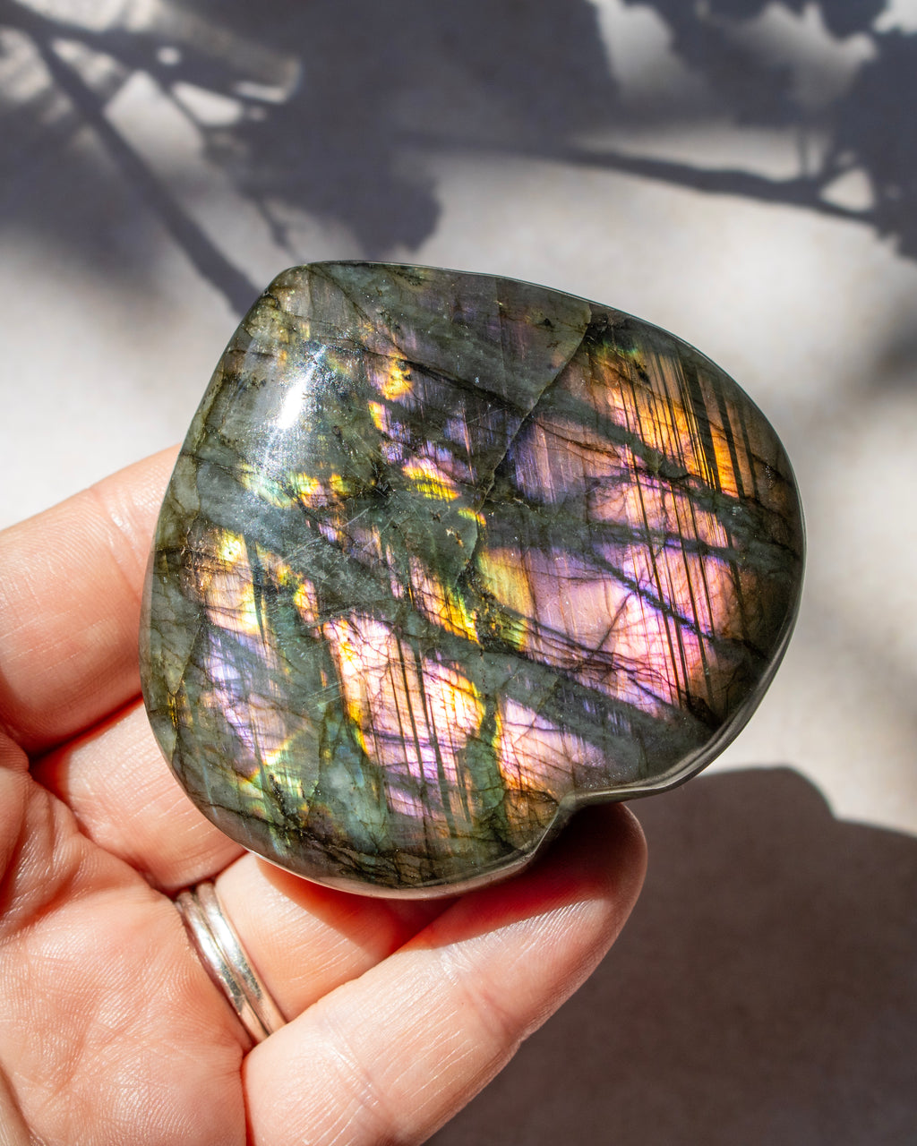 A polished labradorite stone held in a hand, showing a shimmering play-of-color with green, gold, pink, and purple flashes under natural sunlight.