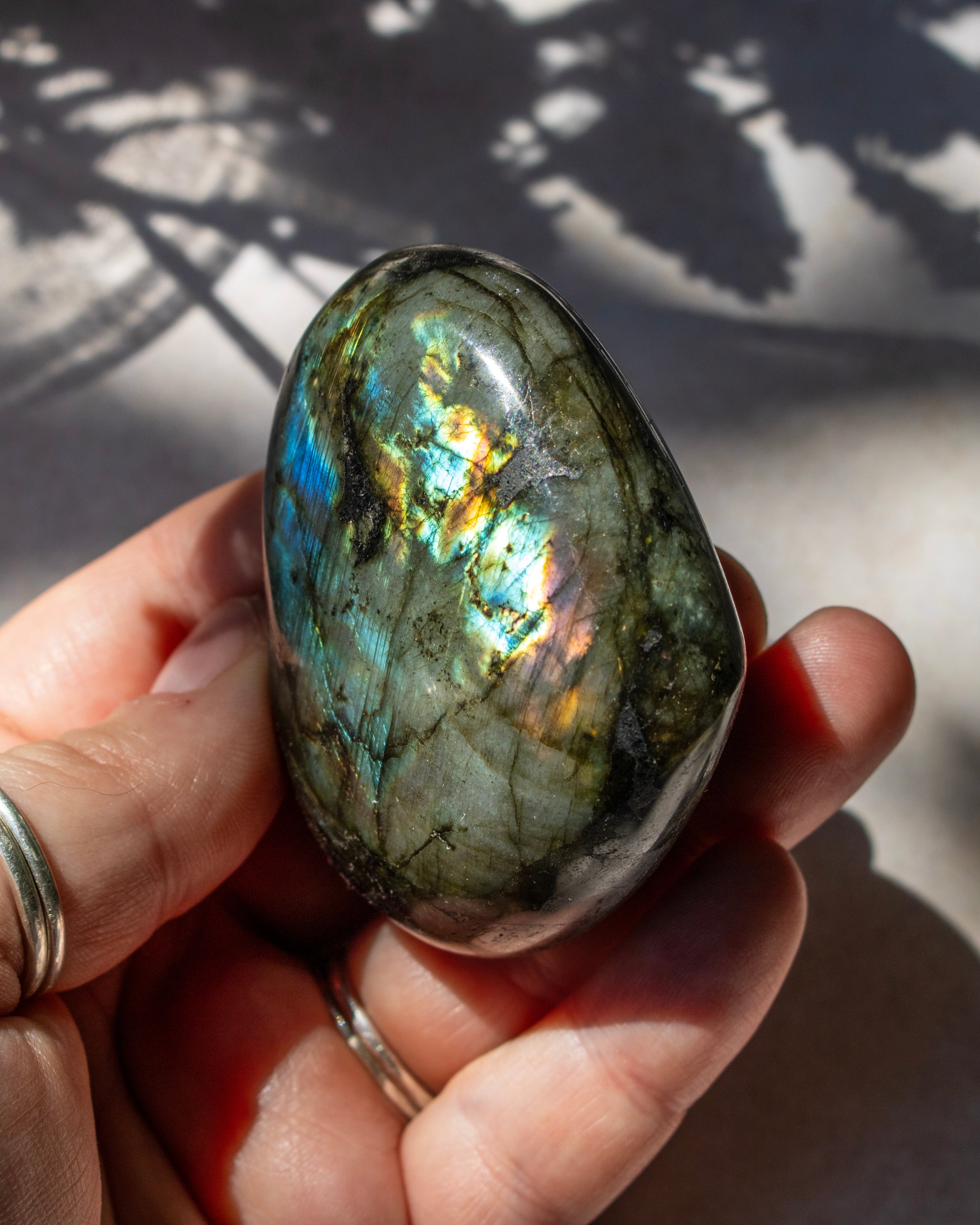 A polished labradorite tumbled stone held in a person's hand, showing vibrant iridescent flashes of blue, green, gold, and purple on a sunlit surface.