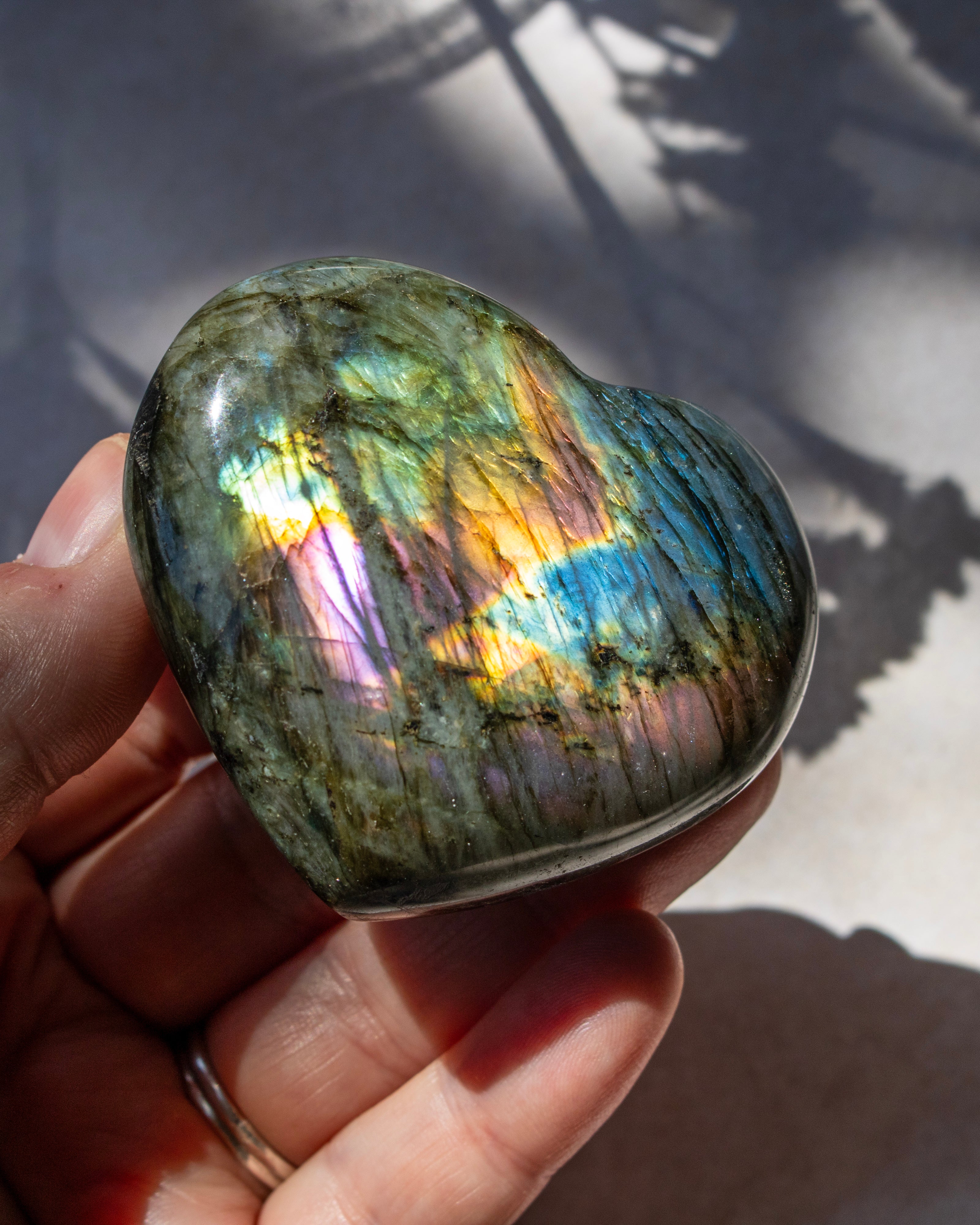 A heart-shaped labradorite crystal held in a person's hand, showcasing its vibrant iridescent rainbow sheen under direct sunlight.
