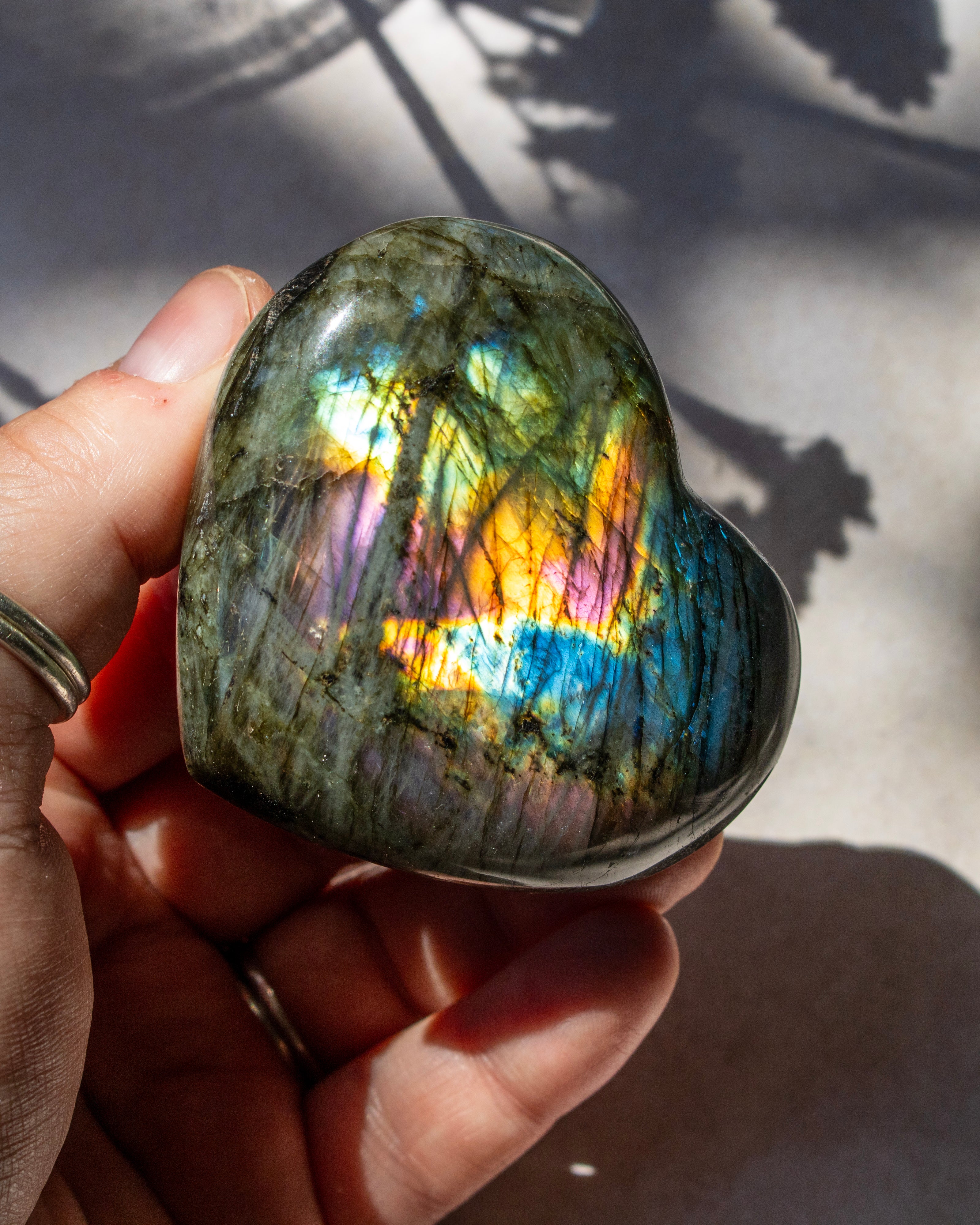 A heart-shaped polished labradorite stone held in a hand, displaying vibrant iridescent rainbow reflections under direct sunlight.