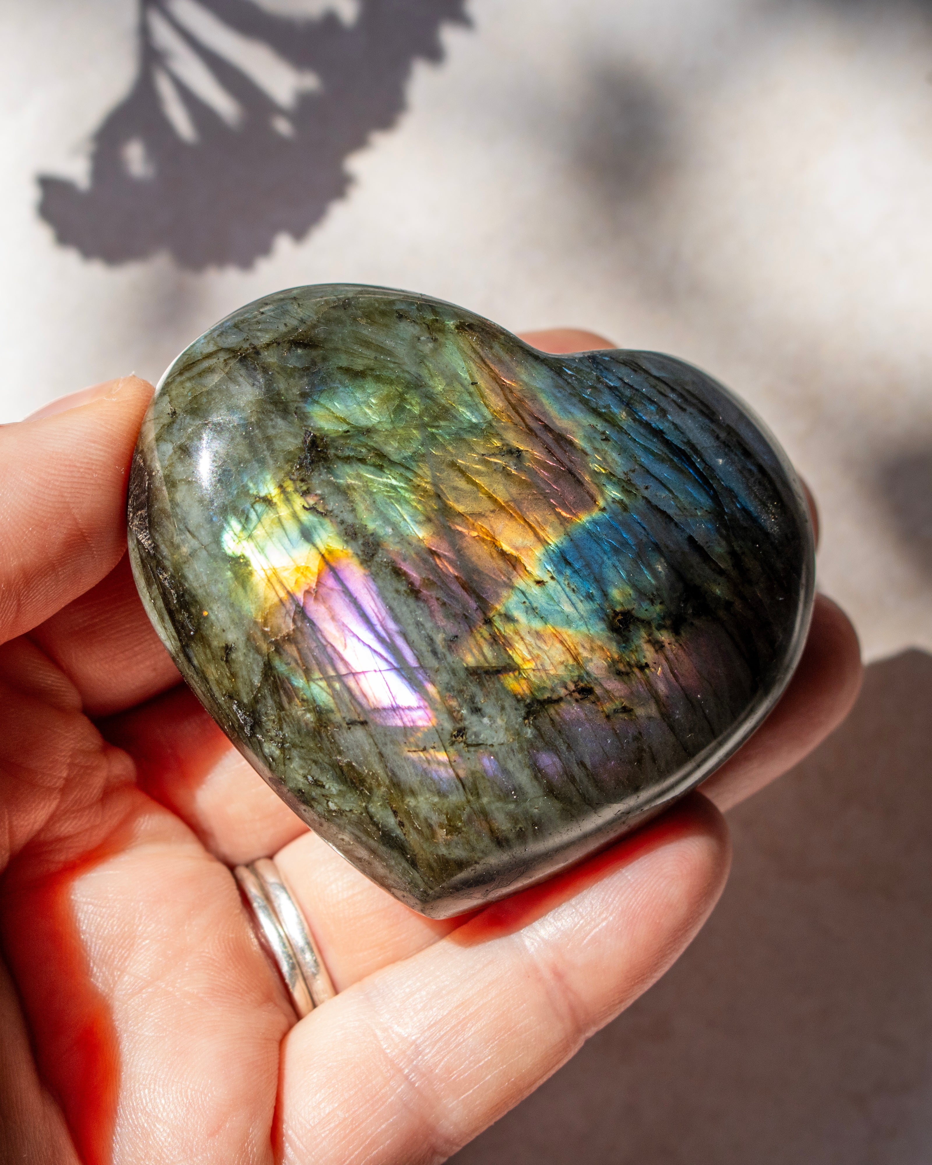 A heart-shaped labradorite crystal held in a person's hand, showing a polished iridescent surface with green, blue, purple, and gold tones under natural sunlight.