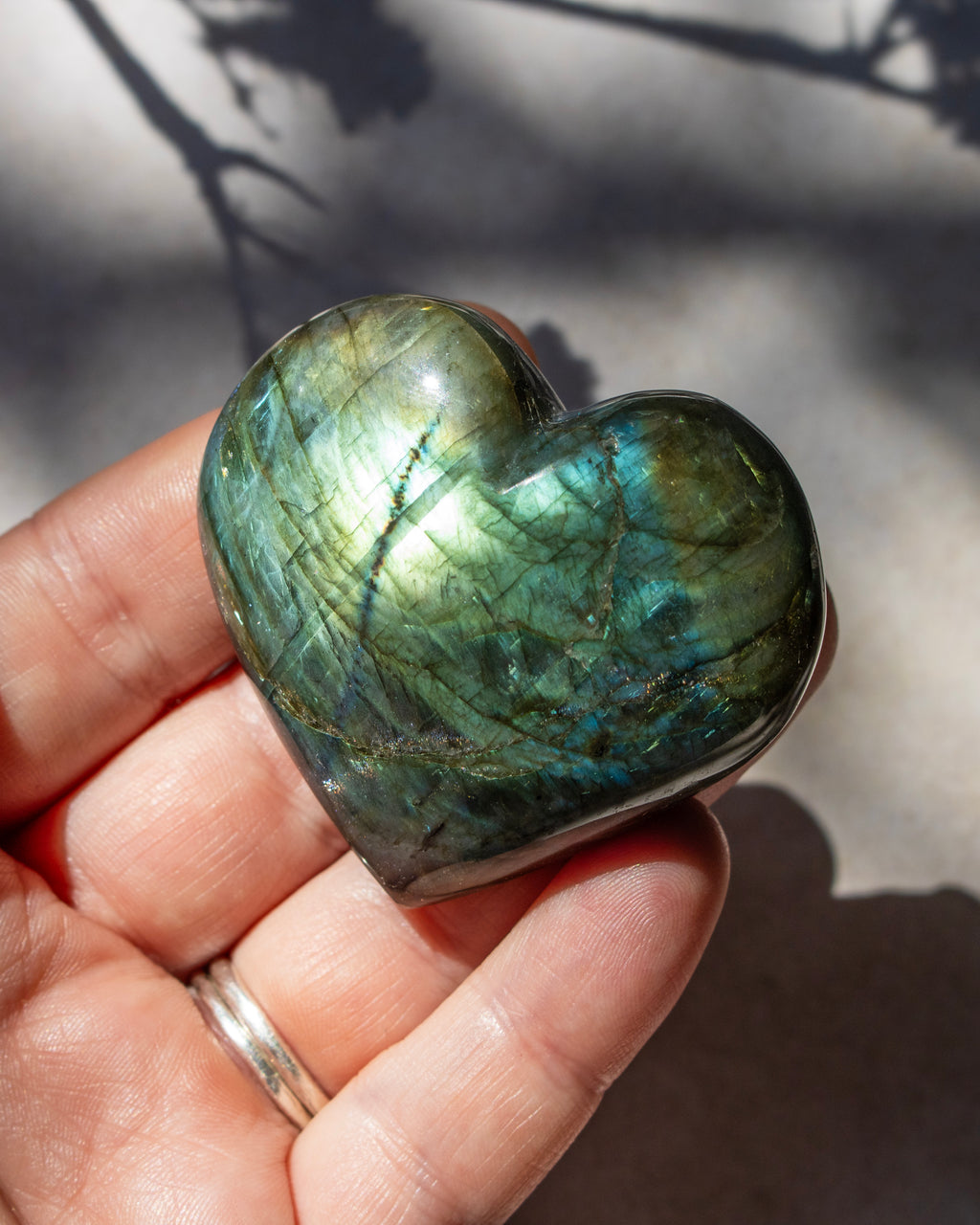 A polished heart-shaped labradorite crystal held in a person's hand, showing shimmering green, blue, and gold iridescence under natural sunlight.