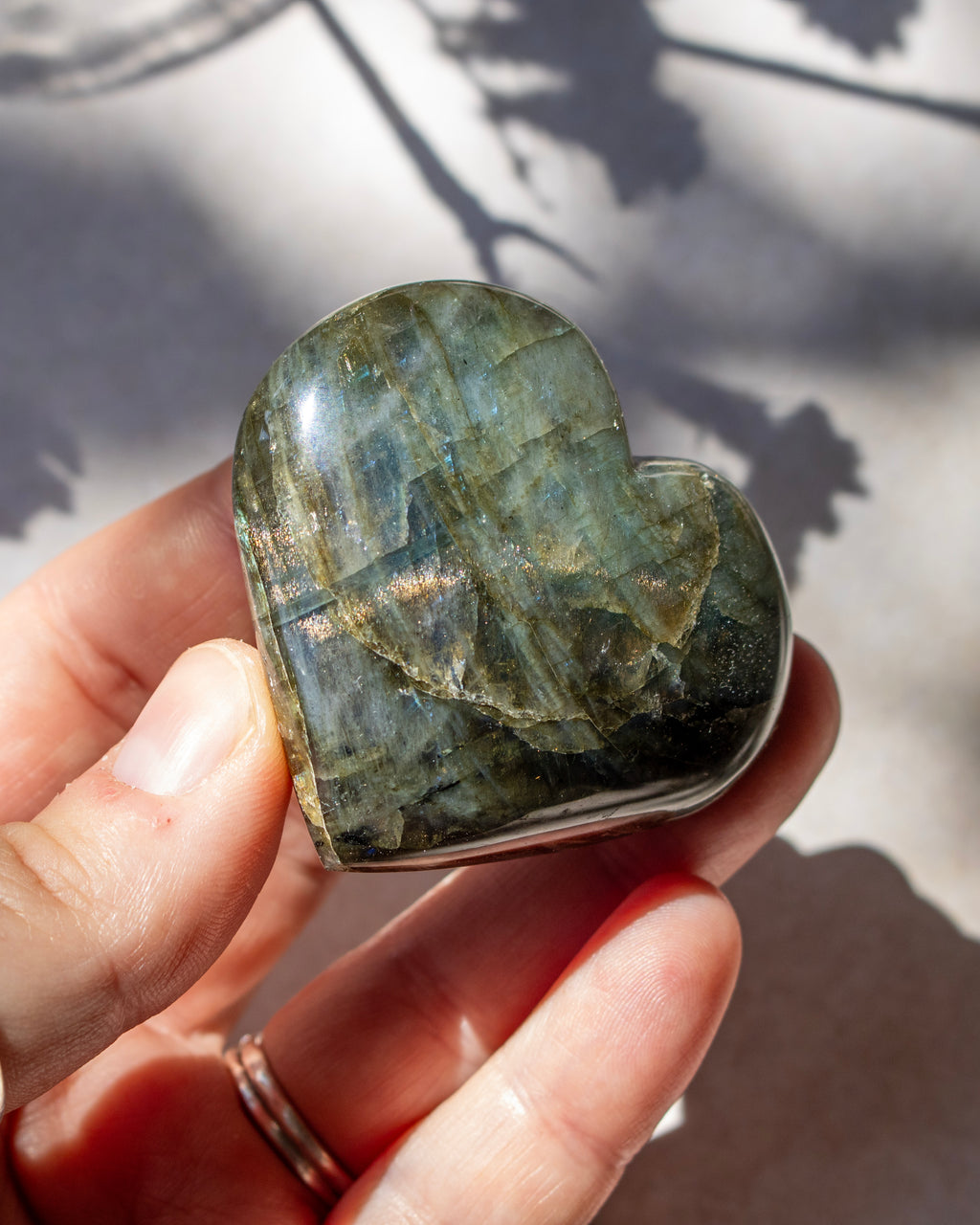 A heart-shaped labradorite gemstone held in a person's hand, showing its characteristic green and gray flashes under natural sunlight with leaf shadows on a light background.