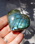 A polished heart-shaped labradorite stone held in a person's hand, showing shimmering blue, green, and gold iridescent flashes under natural sunlight with leaf shadows on the background.