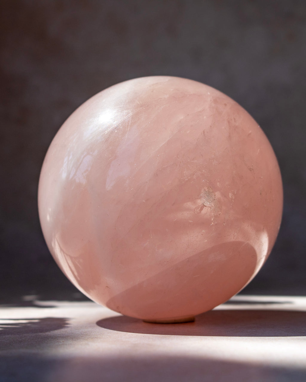 A polished pink rose quartz crystal sphere resting on a dark surface against a dark gray background, with soft sunlight casting a shadow.