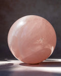A polished pink rose quartz crystal sphere resting on a dark surface against a dark gray background, with soft sunlight casting a shadow.