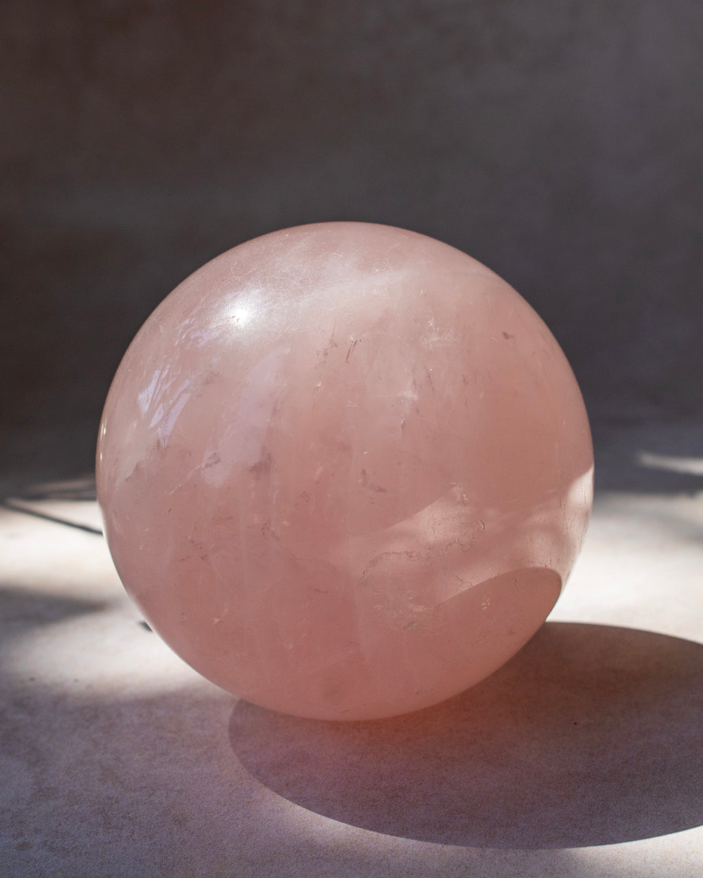 A large smooth pink stone sphere, likely rose quartz, photographed on a dark gray surface with direct sunlight casting a strong shadow.
