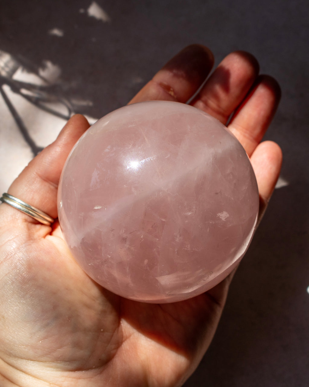 A hand holding a large, smooth, translucent pink rose quartz crystal sphere under natural sunlight.