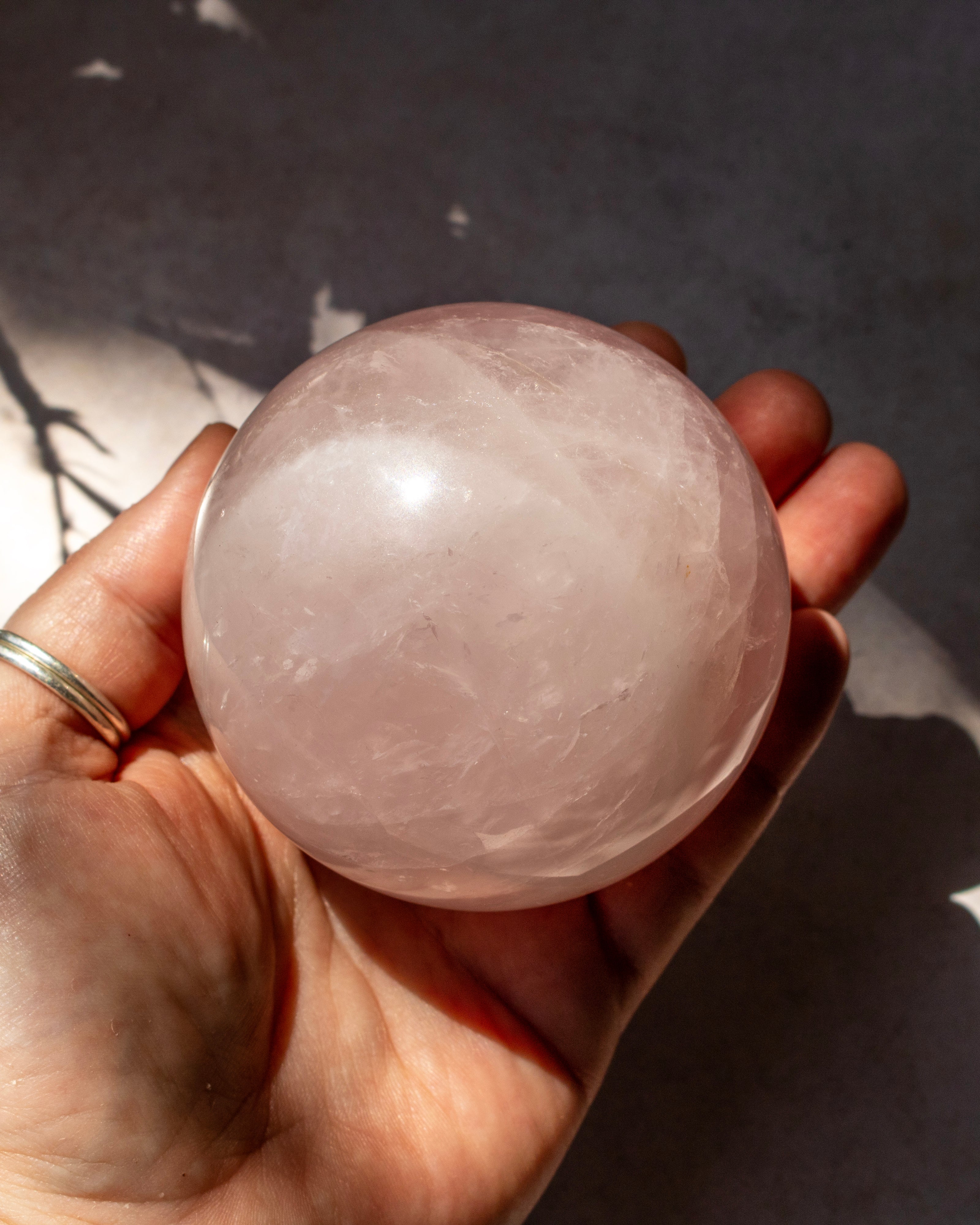 A hand holding a smooth, polished pale pink stone sphere, likely rose quartz, against a dark gray background with soft shadows.