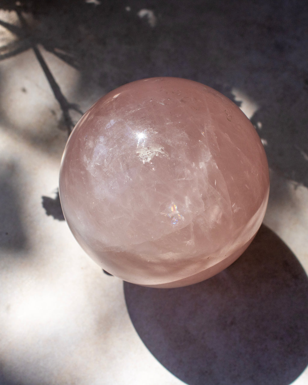 A polished pink quartz crystal sphere resting on a dark surface with dappled sunlight and shadows.
