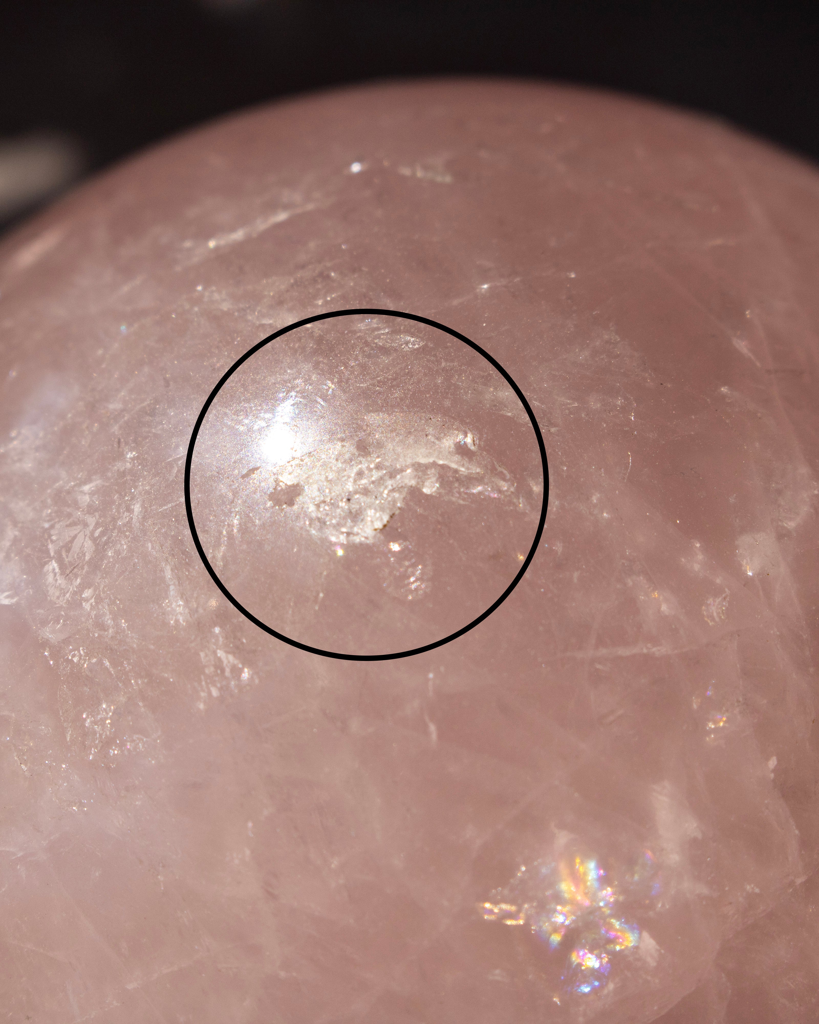 A close-up of a translucent pale pink stone sphere with a rough, textured white area circled in black, showing natural crystalline texture and iridescent highlights.