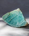A raw amazonite crystal stone with natural rough edges, displaying a light greenish-blue color with white marbling, resting on a light gray surface with a dark gray background.