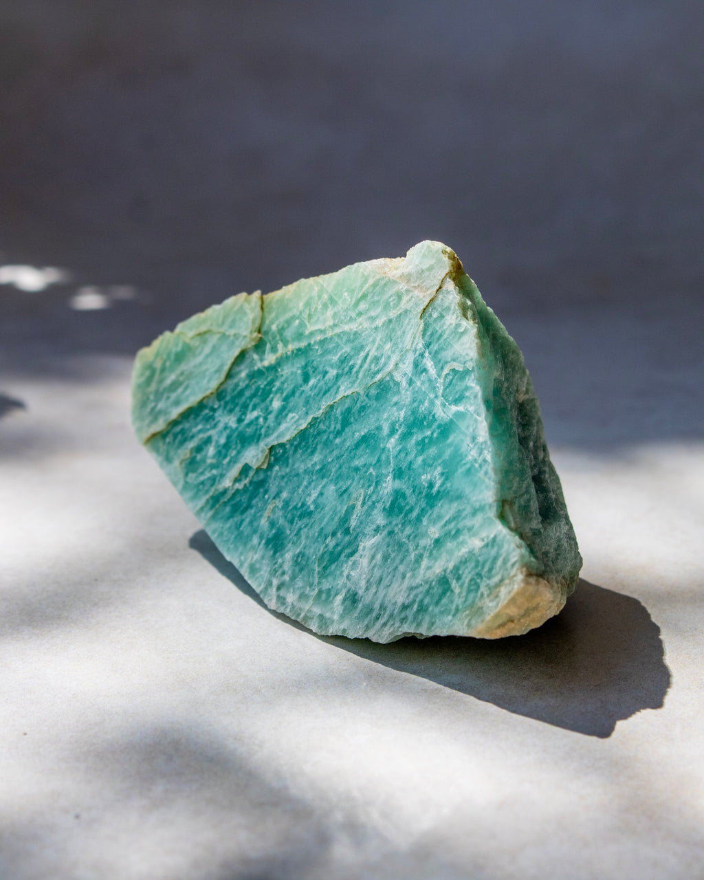 A raw, rough-cut gemstone with a light aqua-green color and white marbling, resting on a light gray surface with soft shadows.