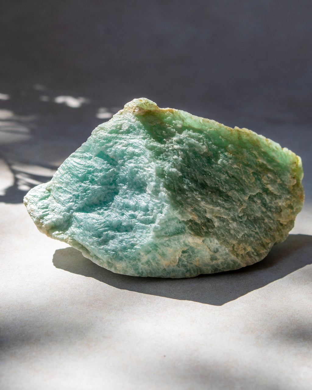 A raw green mineral specimen with a textured, crystalline surface, resting on a neutral gray surface under natural dappled sunlight.