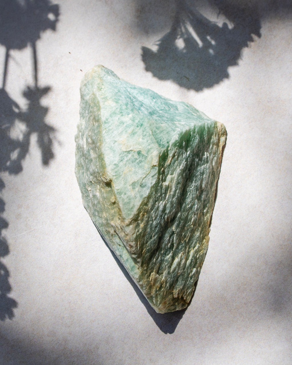 A rough green stone or mineral chunk with natural texture and layered coloration, photographed on a light neutral surface with soft shadows cast across it.