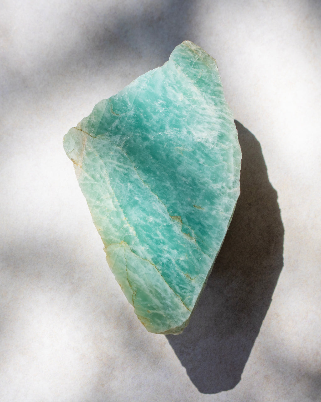 A raw, rough-cut piece of amazonite gemstone with natural green and white marbling, casting a sharp shadow on a textured white surface.