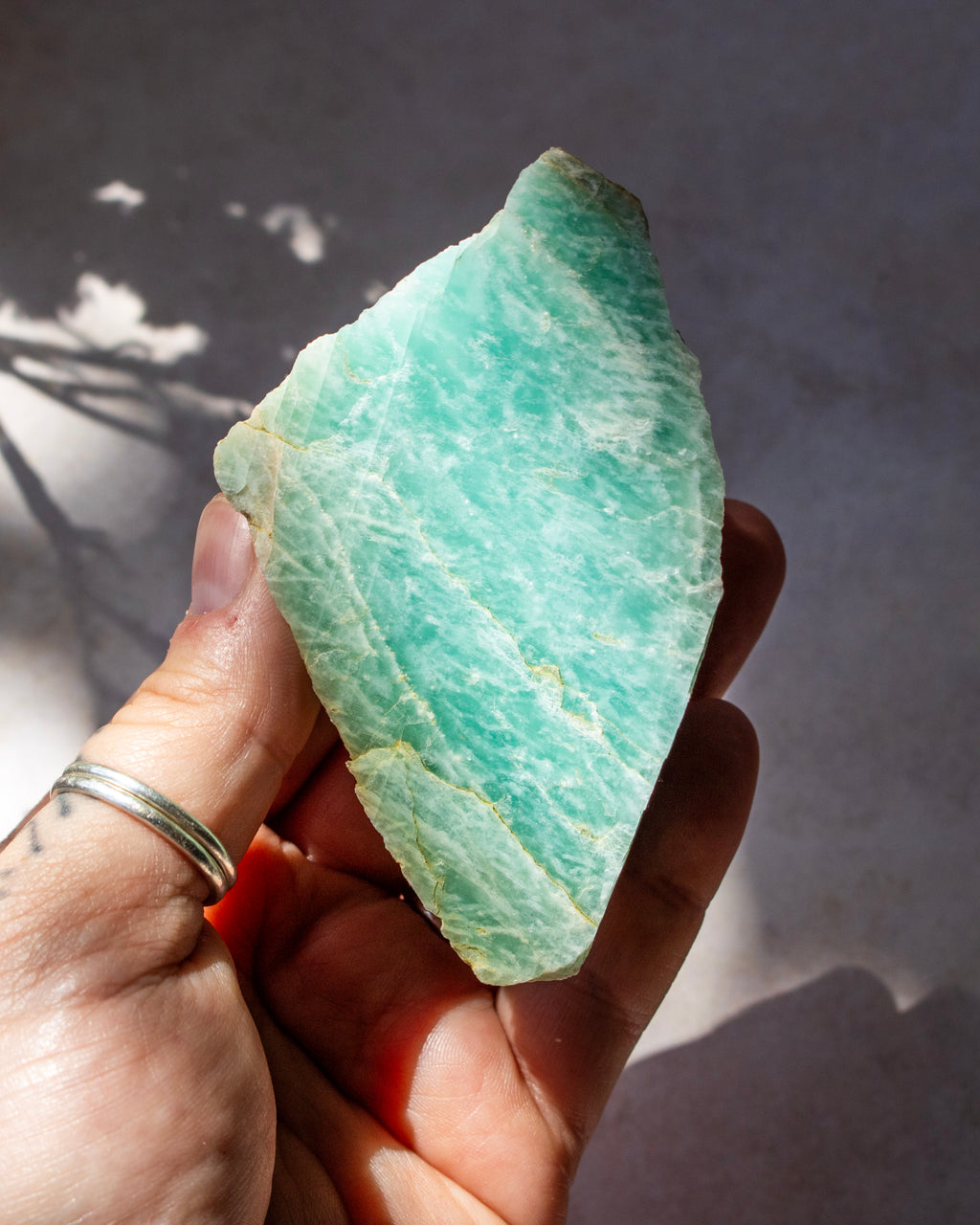A hand holding a raw, rough green aventurine crystal stone with natural striations, set against a neutral gray background with soft shadows.