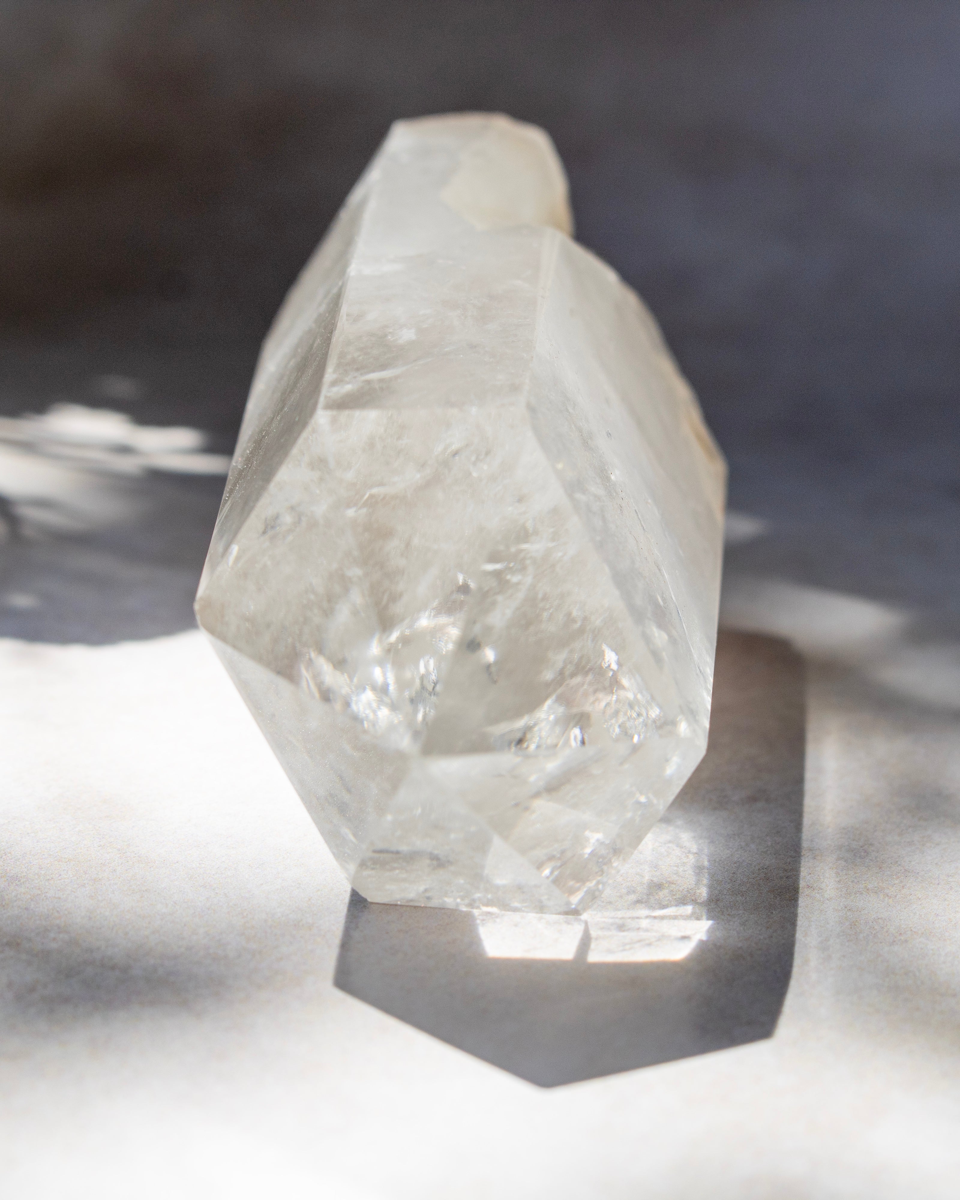 A natural clear quartz crystal cluster with geometric facets, standing upright on a reflective surface with dramatic sunlight and shadows.
