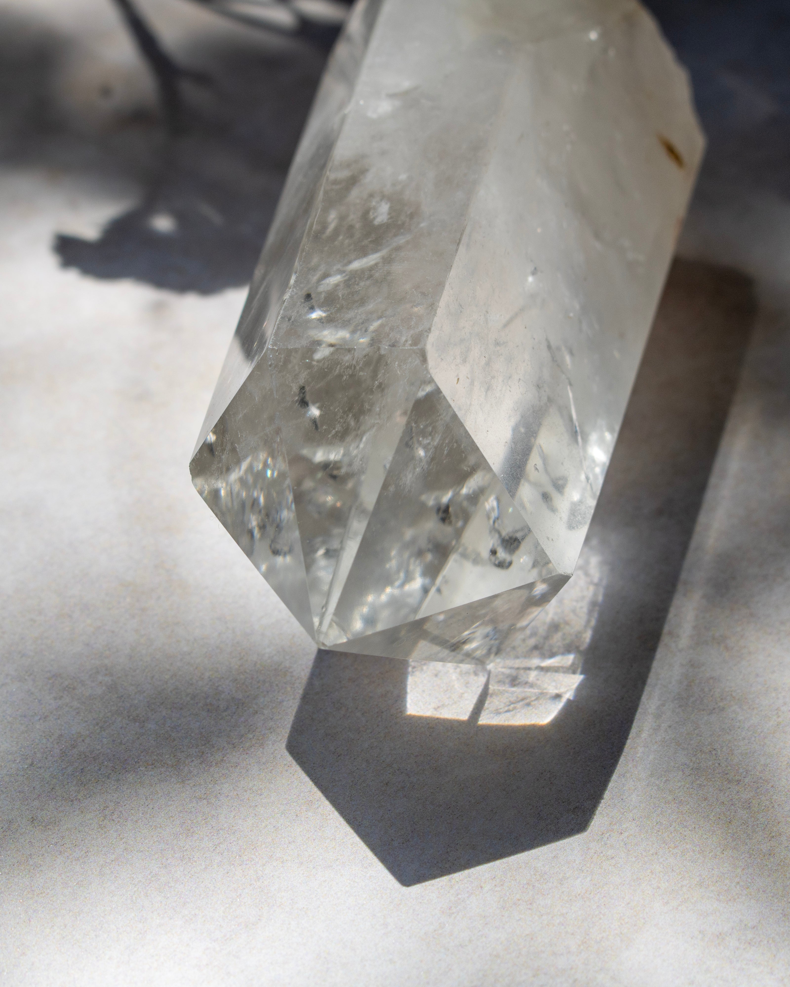 A clear faceted crystal point with natural inclusions, photographed on a light surface with strong sunlight and shadows.