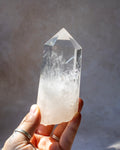 A hand holding a clear quartz crystal point with natural white and cloudy inclusions, set against a neutral gray background.