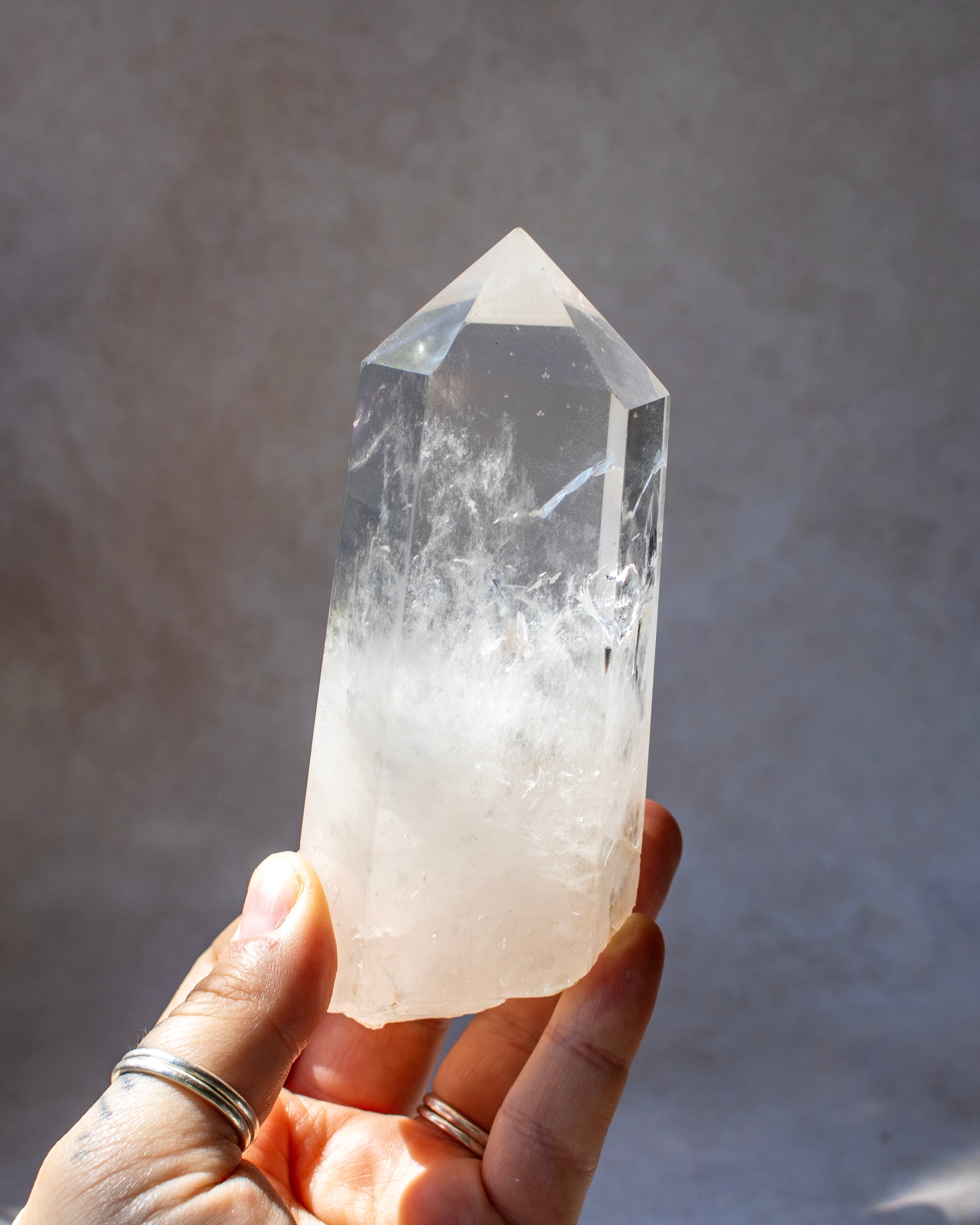 A hand holding a clear quartz crystal point with natural white and cloudy inclusions, set against a neutral gray background.