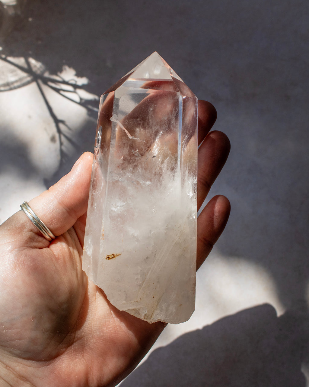 A hand holding a large, clear quartz point crystal with natural inclusions, set against a gray concrete background with dappled sunlight and shadows.