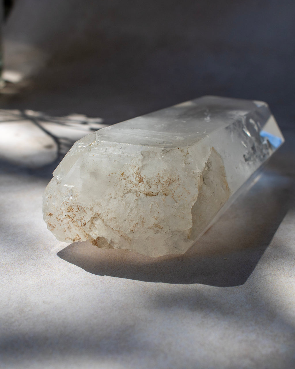 A raw quartz crystal point with rough, textured off-white and light tan matrix, resting on a light gray surface under dramatic, dappled sunlight and shadows.