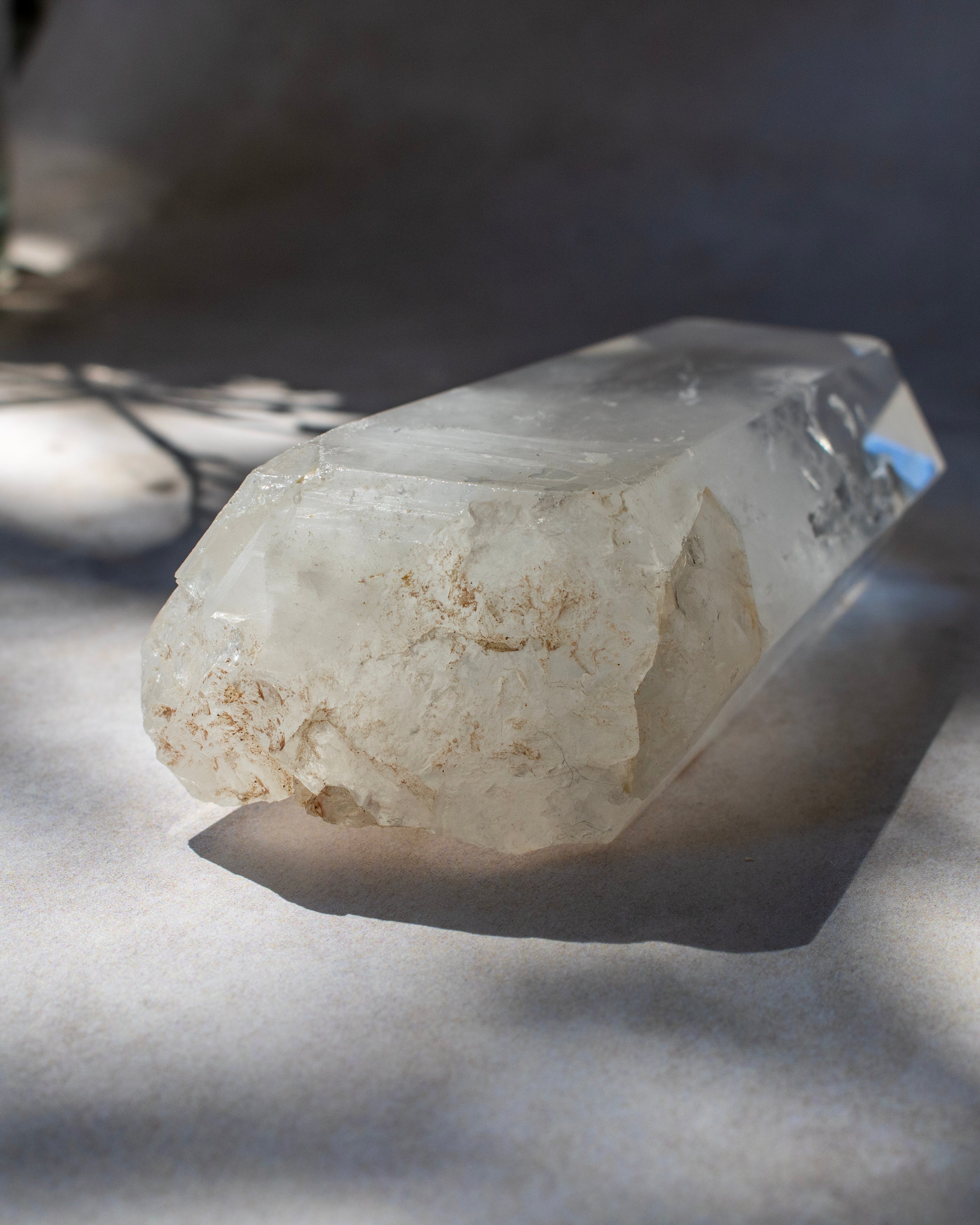 A raw quartz crystal point with rough, textured off-white and light tan matrix, resting on a light gray surface under dramatic, dappled sunlight and shadows.