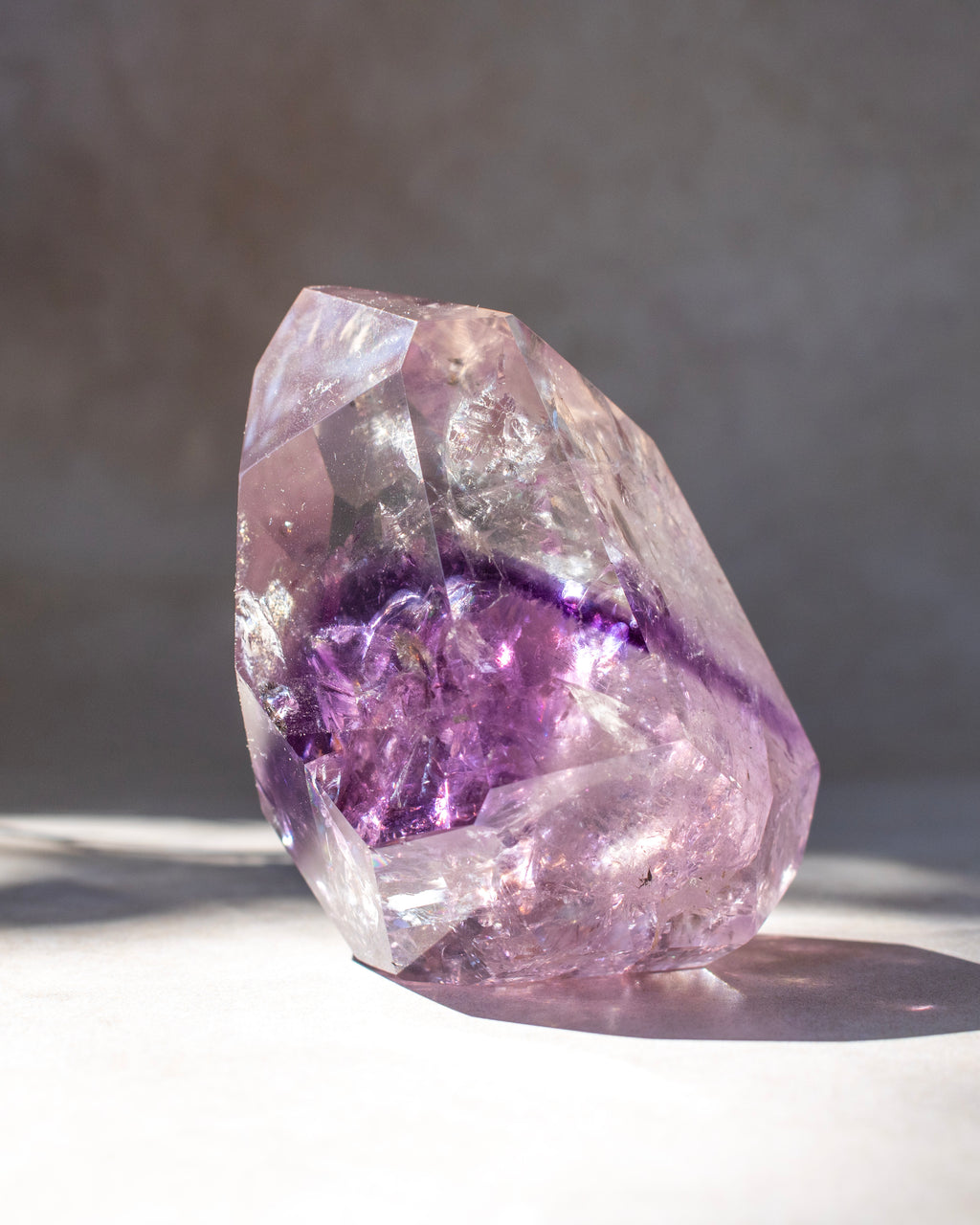 A natural amethyst crystal cluster with a prominent purple band around the middle, photographed on a light surface with a soft gray background.