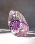 A natural amethyst crystal cluster with a prominent purple band around the middle, photographed on a light surface with a soft gray background.